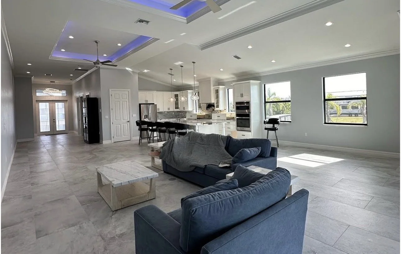Interior of a modern, spacious living area with large windows overlooking water, a blue accent wall, a dining table with chairs, and doors leading to an outdoor space.
