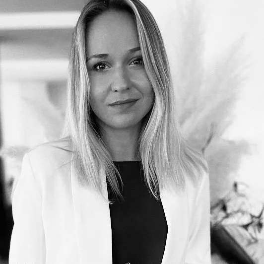 A woman with shoulder-length blonde hair, wearing a white blazer over a dark top, smiling softly in a black-and-white photo.