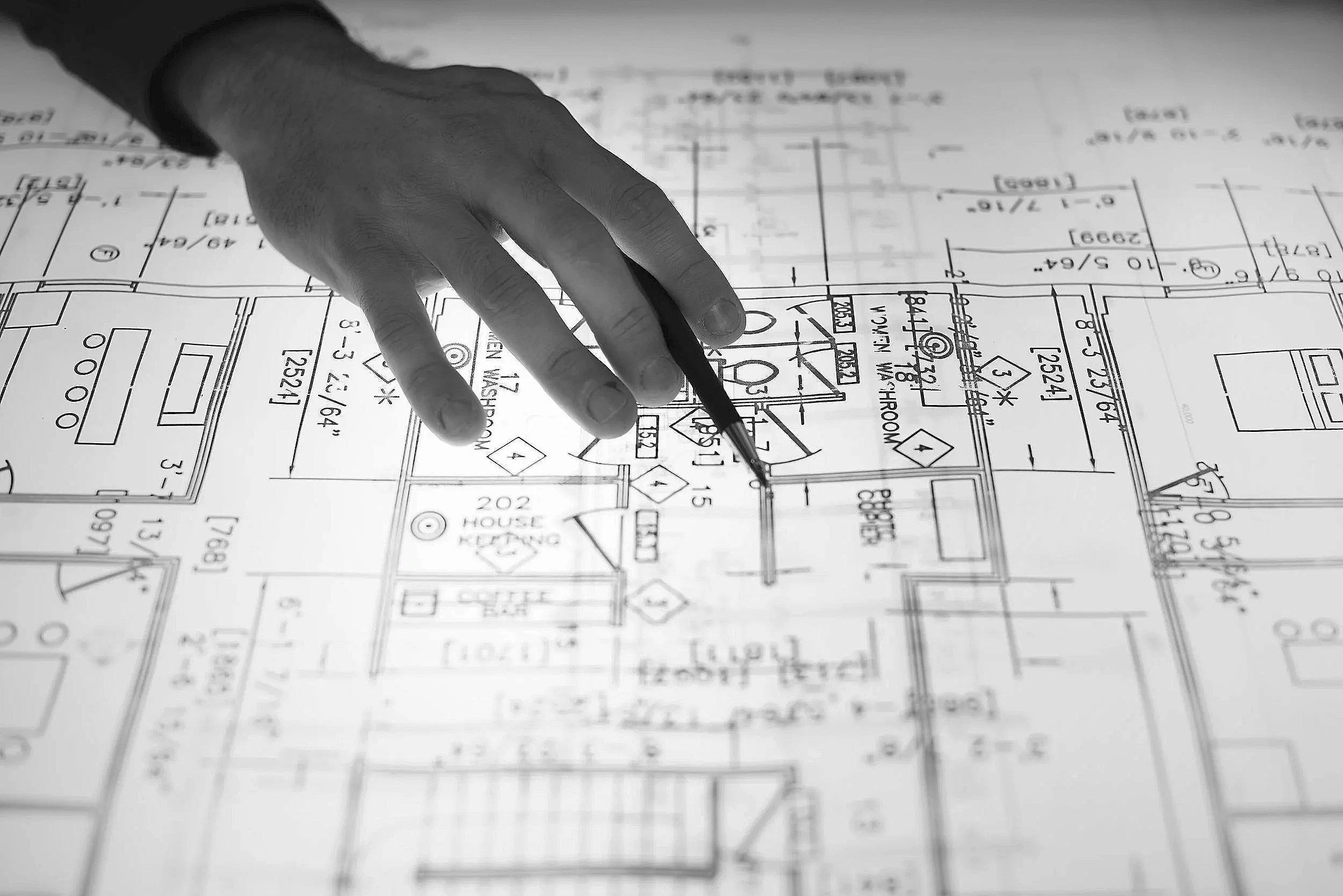 A hand holding a pencil pointing at a detailed architectural blueprint or floor plan on paper.