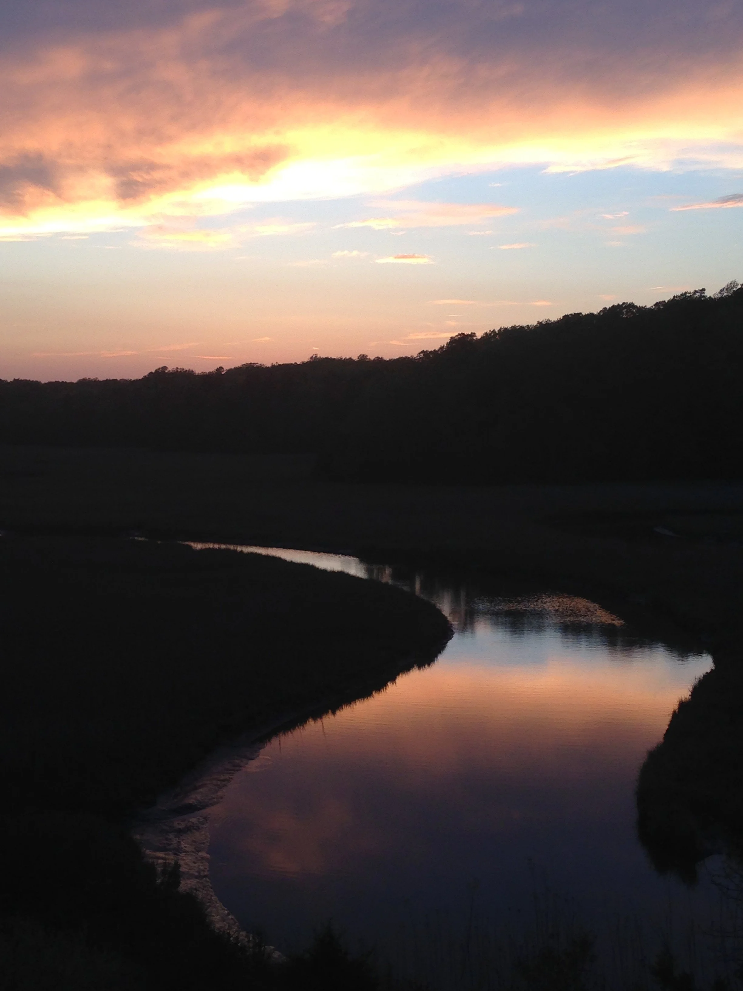 Sunset over a winding river with a dark silhouetted hillside and colorful sky with pink, orange, and blue hues.