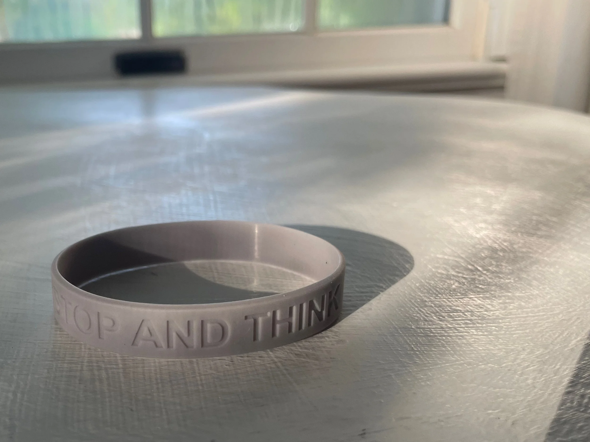 A grey silicone wristband with the text 'STOP AND THINK' embossed around it, placed on a textured white surface near a window.