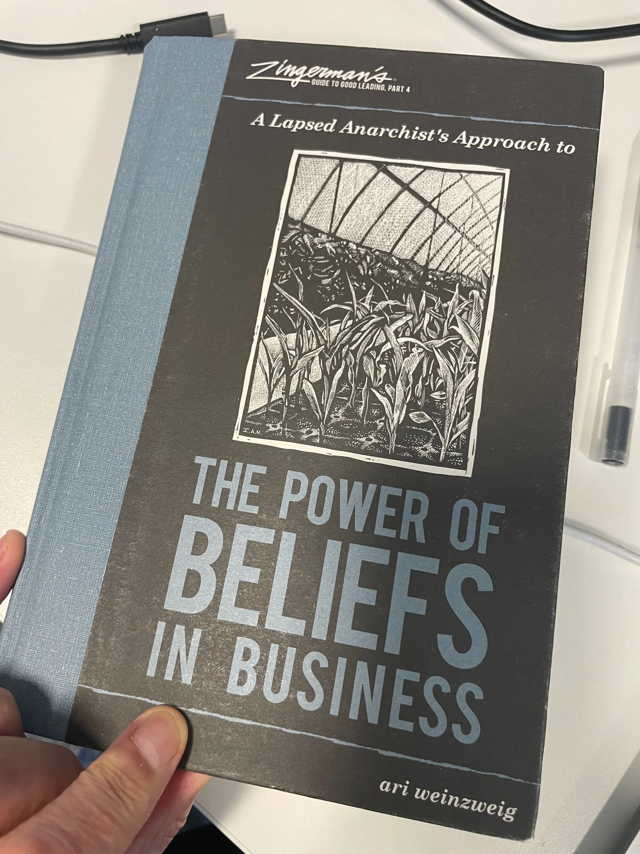 Book titled 'The Power of Beliefs in Business' by Ari Weinzweig, part of Zingerman's Guide to Good Leadership, Part 4, with a cover illustration of a greenhouse or garden with plants.