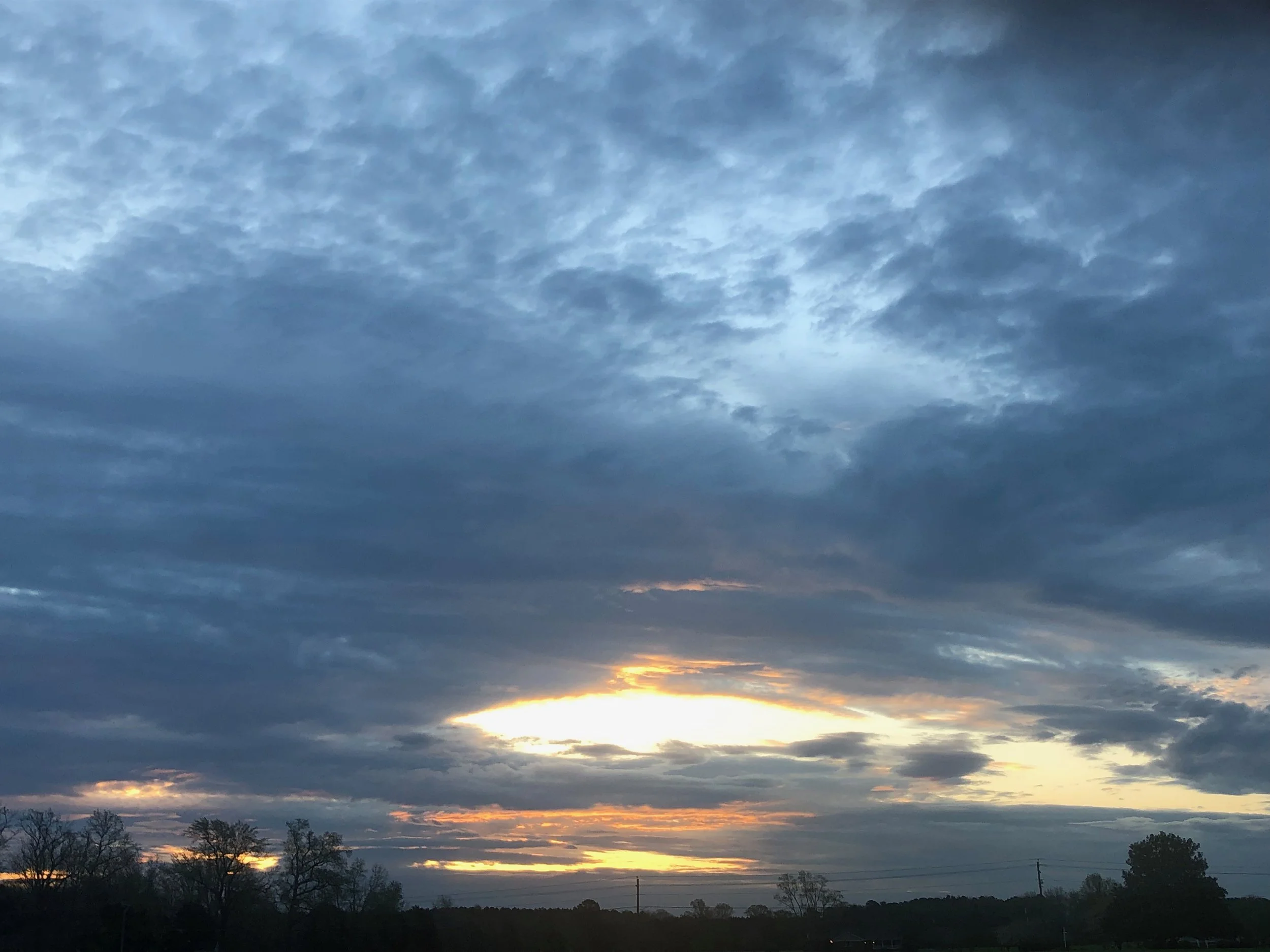 Overcast sky at sunset with dark clouds and a small clearing of sunlight near the horizon, with silhouettes of trees and power lines in the foreground.