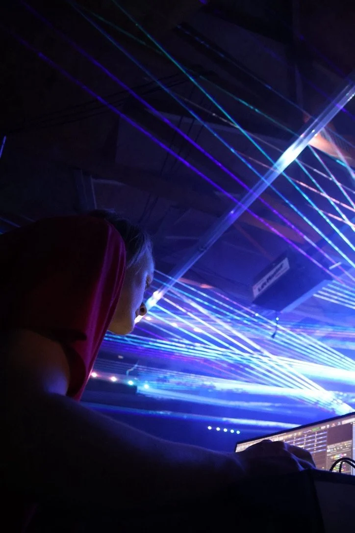 A person is operating DJ equipment at a nightclub or event, with colorful laser lights in the background creating a vibrant atmosphere.