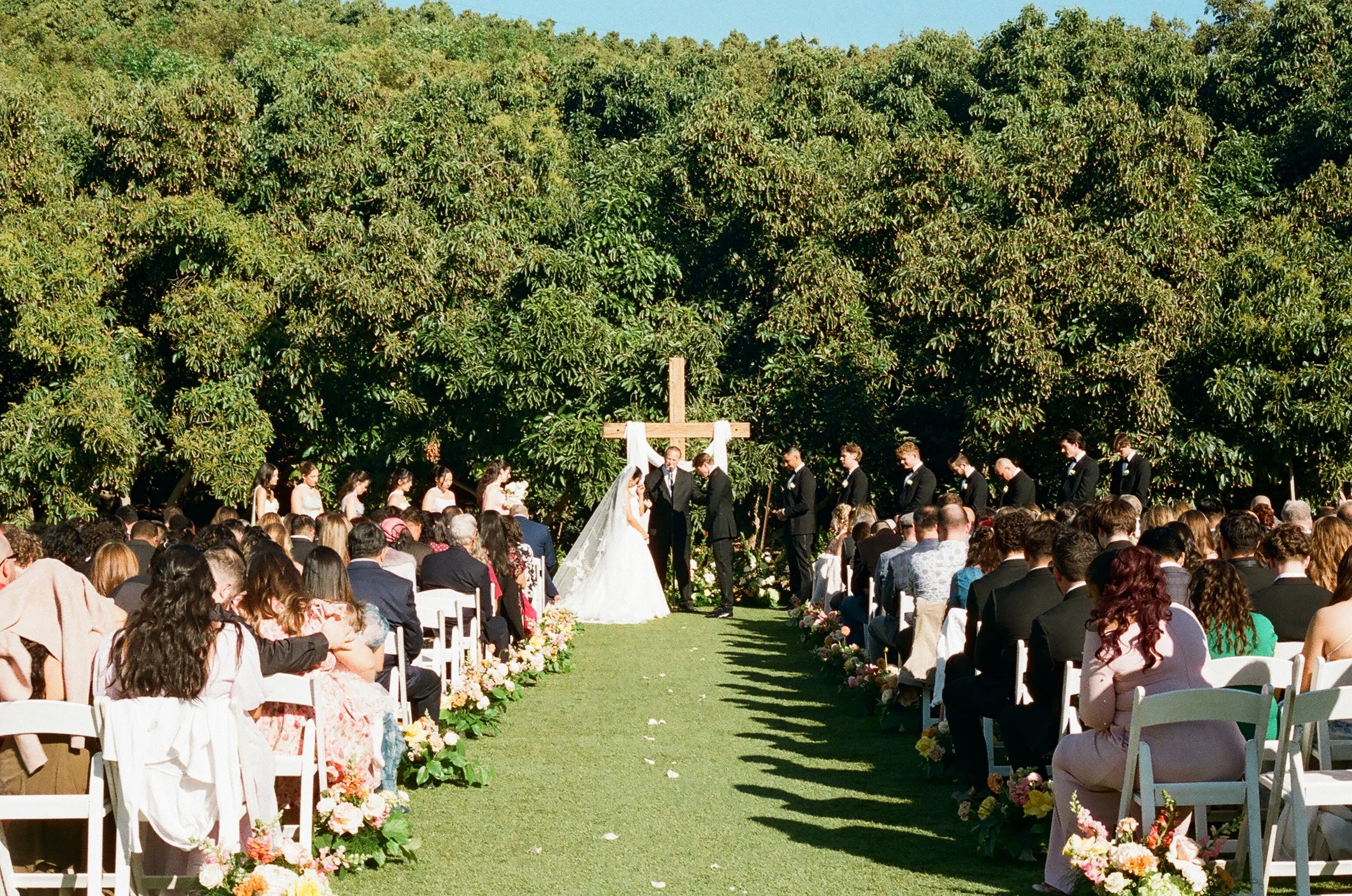 Outdoor wedding ceremony with a bride and groom standing under a cross, surrounded by a large congregation, with lush green trees and floral decorations.