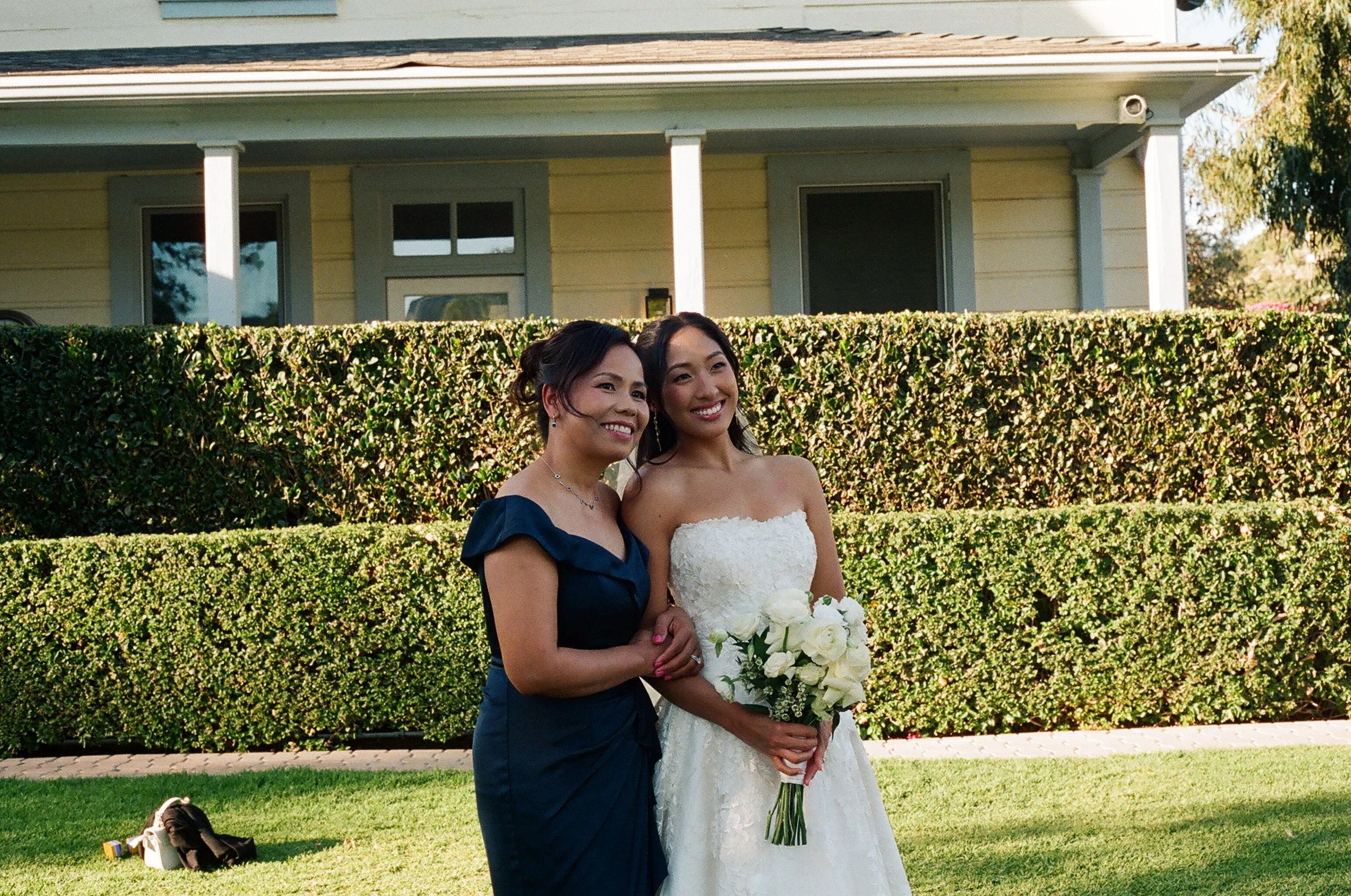 Two women, one in a dark blue dress and the other in a white wedding gown, standing together outdoors with a green hedge and a house in the background, smiling at the camera. The woman in white is holding a bouquet of white roses.