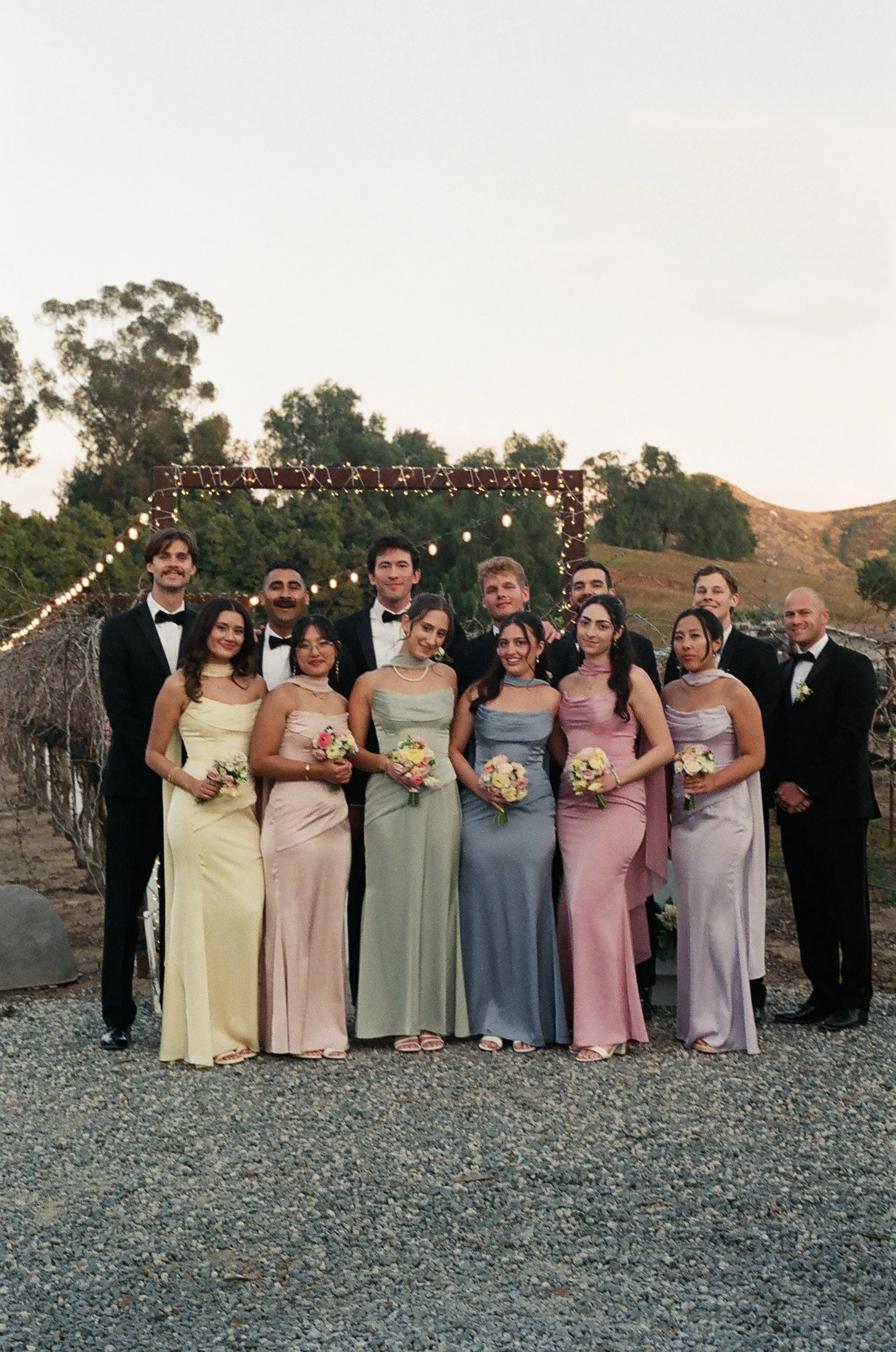A group of mostly young adults in formal attire, with some women holding bouquets of flowers, standing outdoors at sunset with a scenic hillside and trees in the background, decorated with string lights.