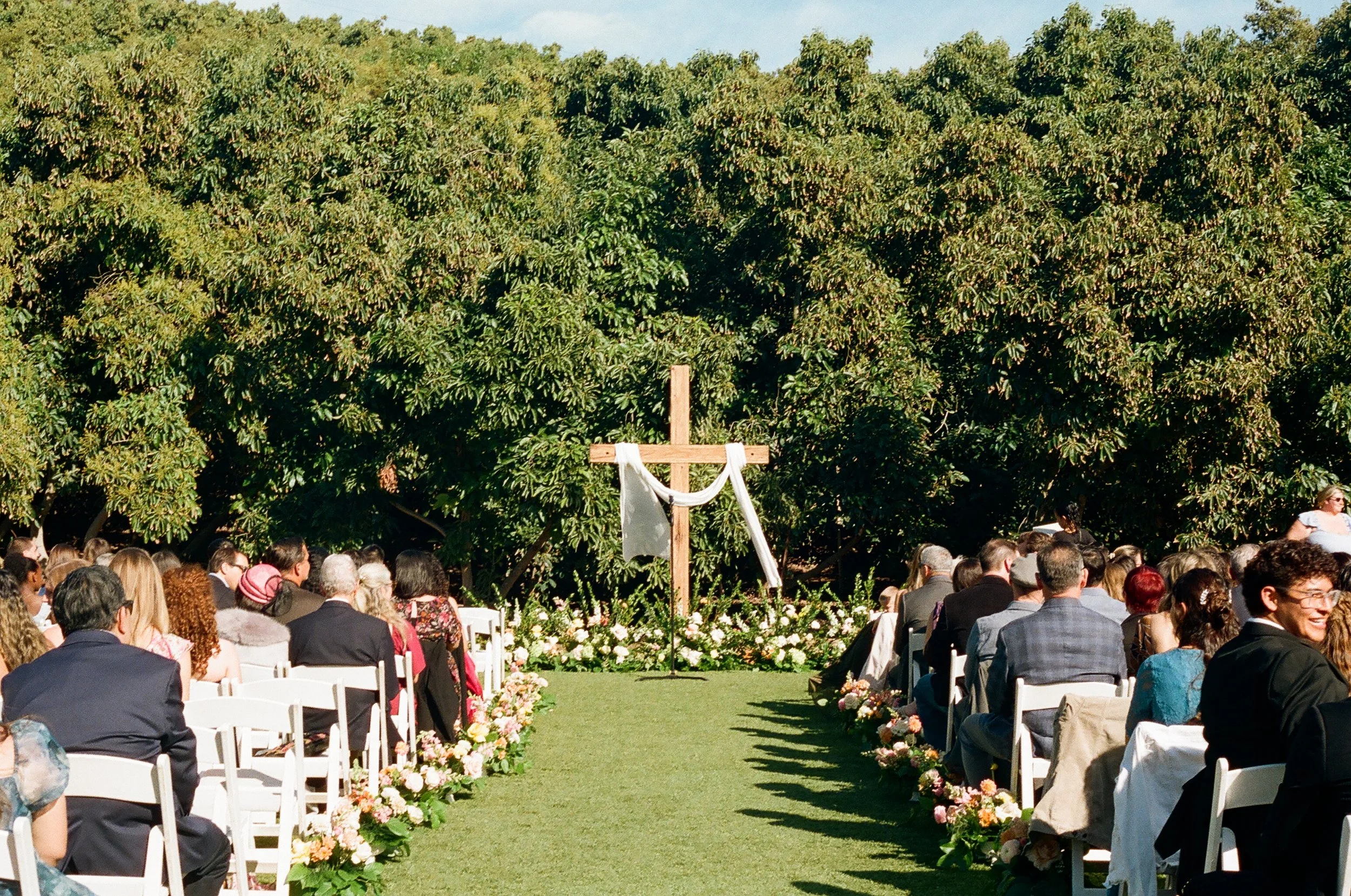 Outdoor wedding ceremony with guests seated on either side of a green aisle decorated with flowers, under large trees with lush greenery.