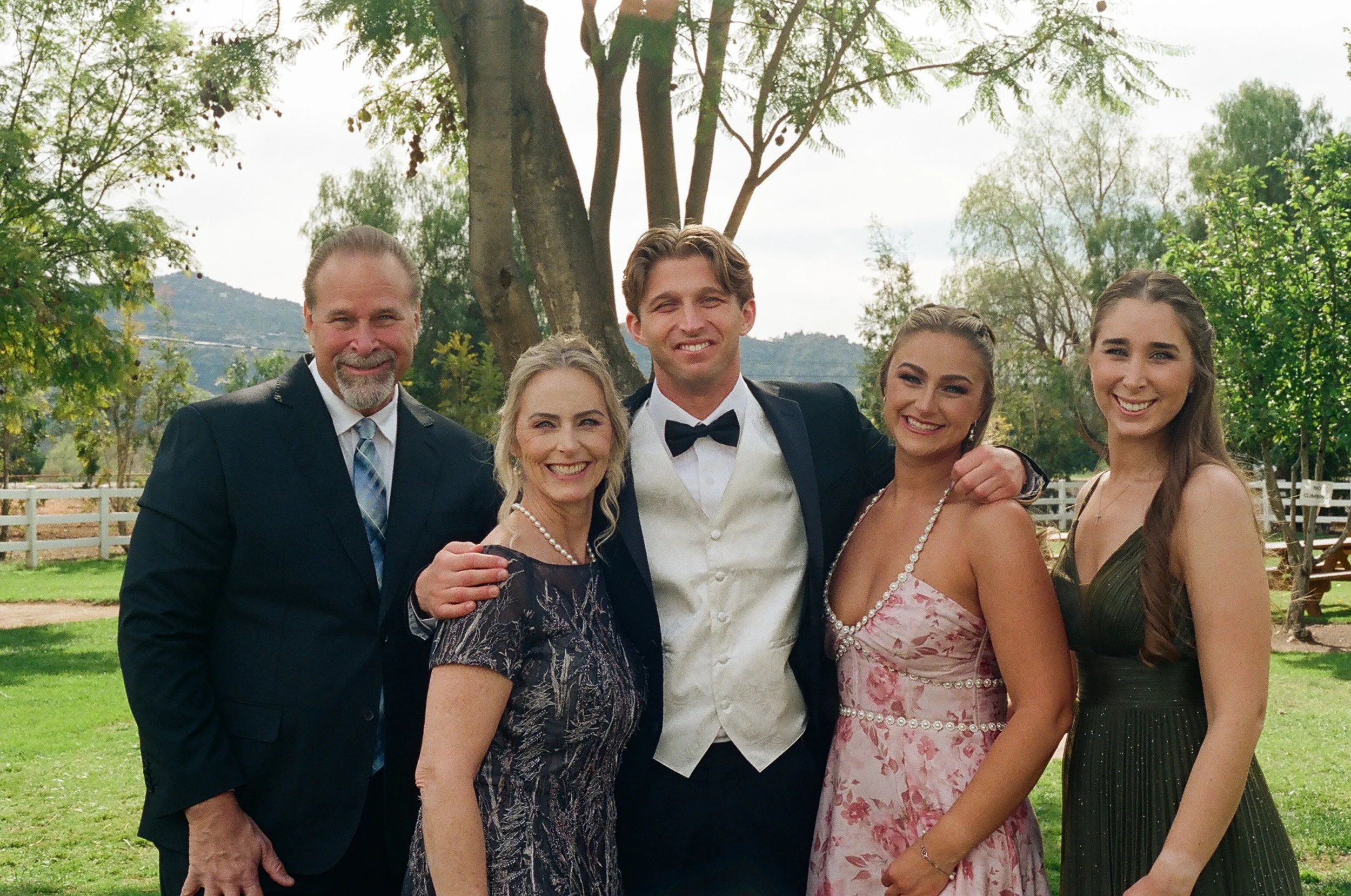 A group of six people, three men and three women, dressed in formal attire, standing outdoors in a park-like setting with trees and a white fence in the background. They are smiling and posing for a photo, possibly at a wedding or special event.