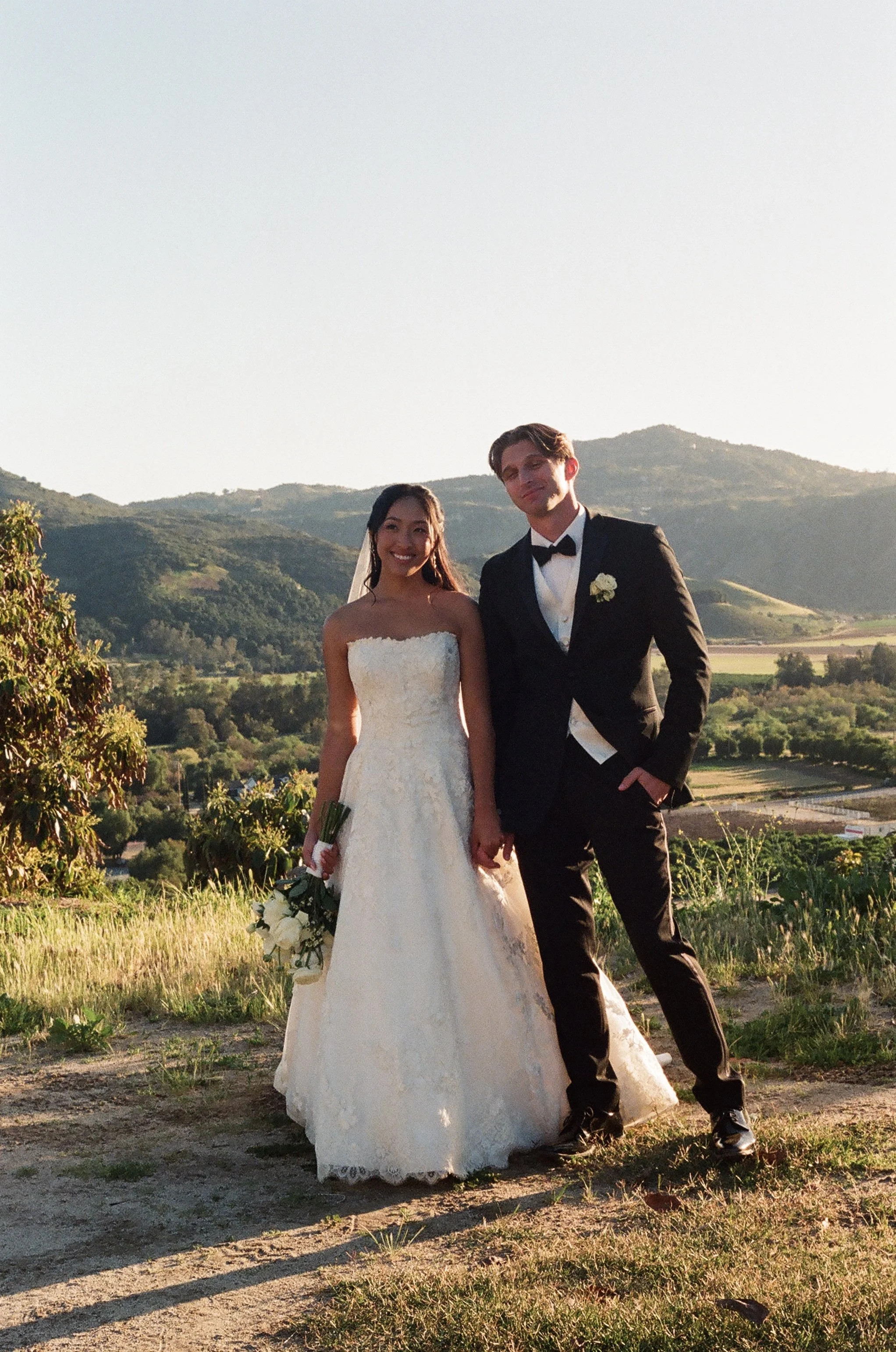 A bride and groom are standing outdoors on a hillside with mountains in the background during sunset. The bride is wearing a strapless white wedding gown and holding a bouquet of white roses, while the groom is dressed in a black tuxedo with a bow ti