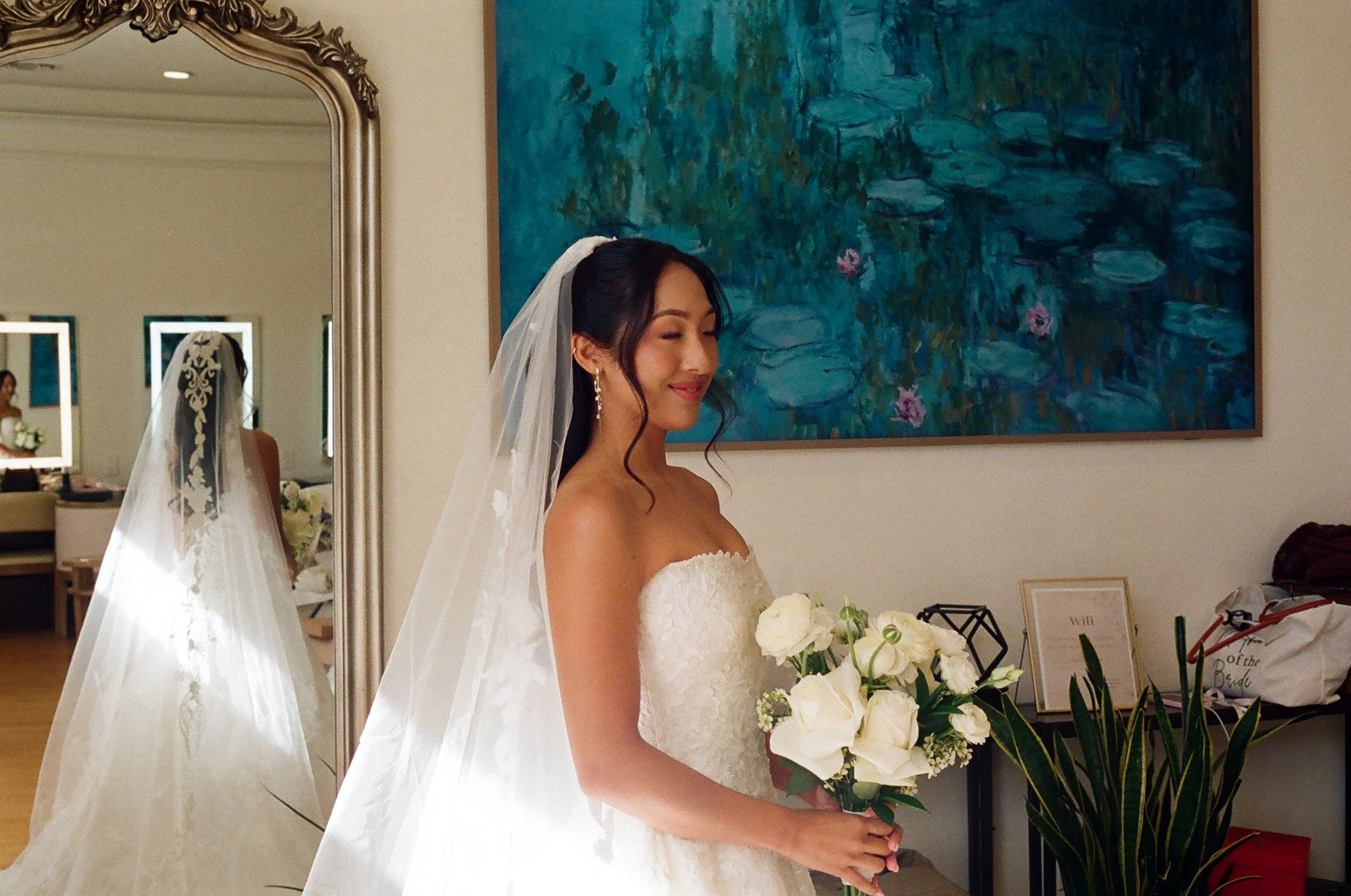 A bride in a white wedding dress holding a bouquet of white flowers, standing in front of a large painting of water lilies, with her eyes closed and a gentle smile on her face.
