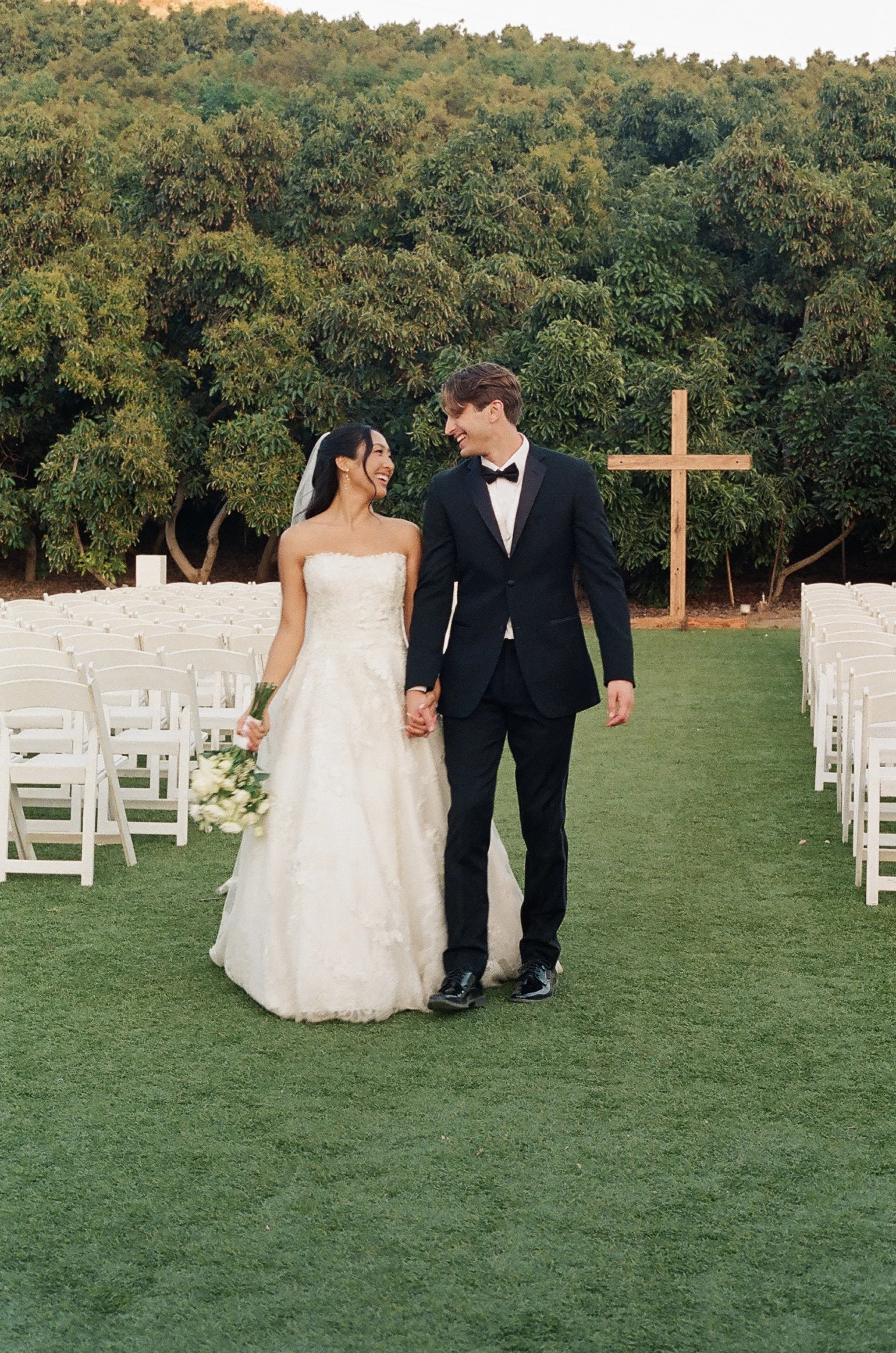 A bride and groom holding hands and smiling at each other during an outdoor wedding ceremony, with rows of white chairs and a large wooden cross in the background.