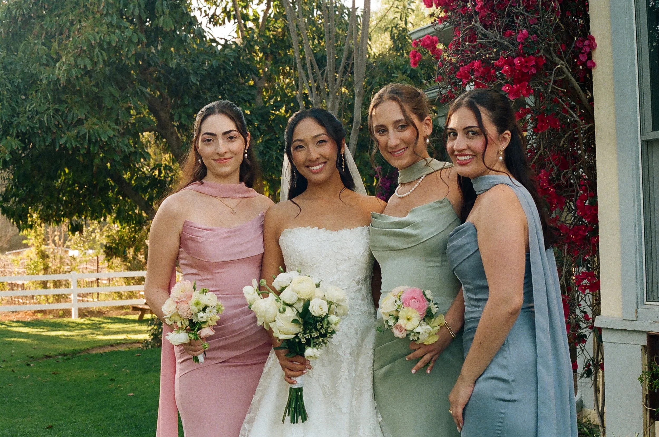 A bride in a white wedding dress with a bouquet standing between four women in pastel-colored dresses, holding bouquets, outdoors with trees and a flowering bush in the background.