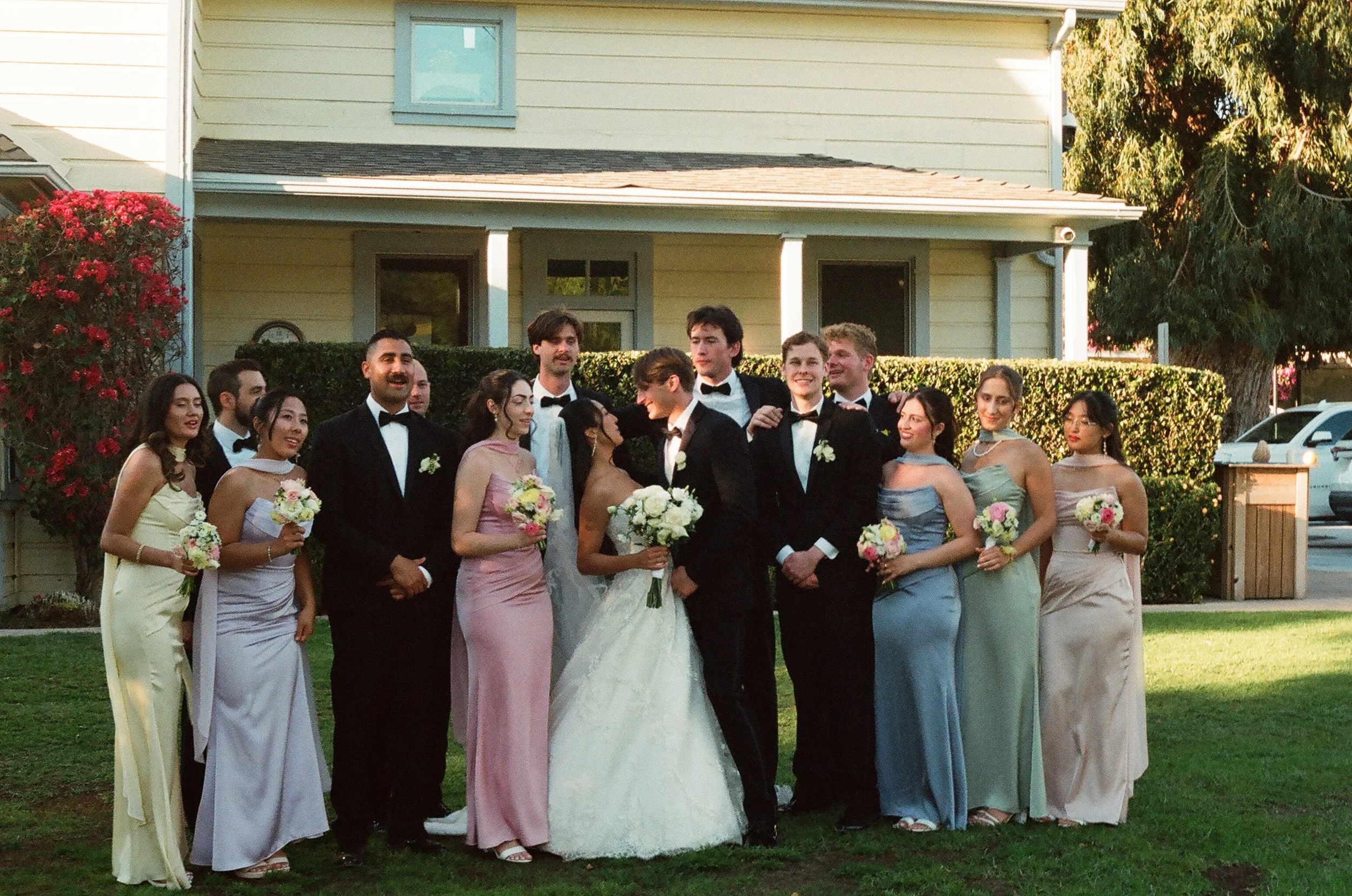 A group of people dressed in formal attire, including tuxedos and evening gowns, standing together outdoors in front of a house with a hedge and flowering bush, celebrating a wedding.