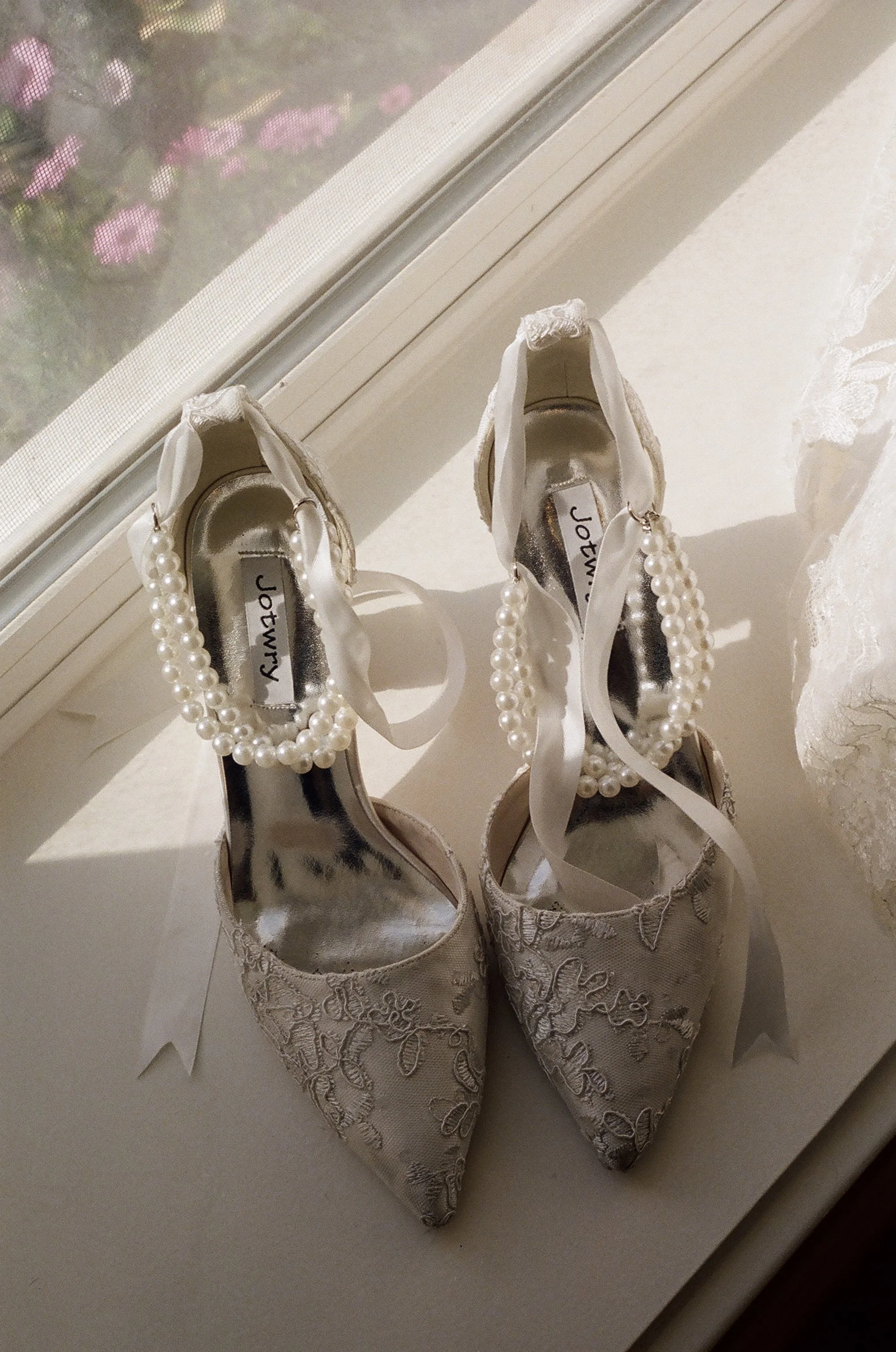 A pair of white lace high heels with pearl ankle straps resting on a white surface near a window with lace curtains, with pink and purple flowers outside.