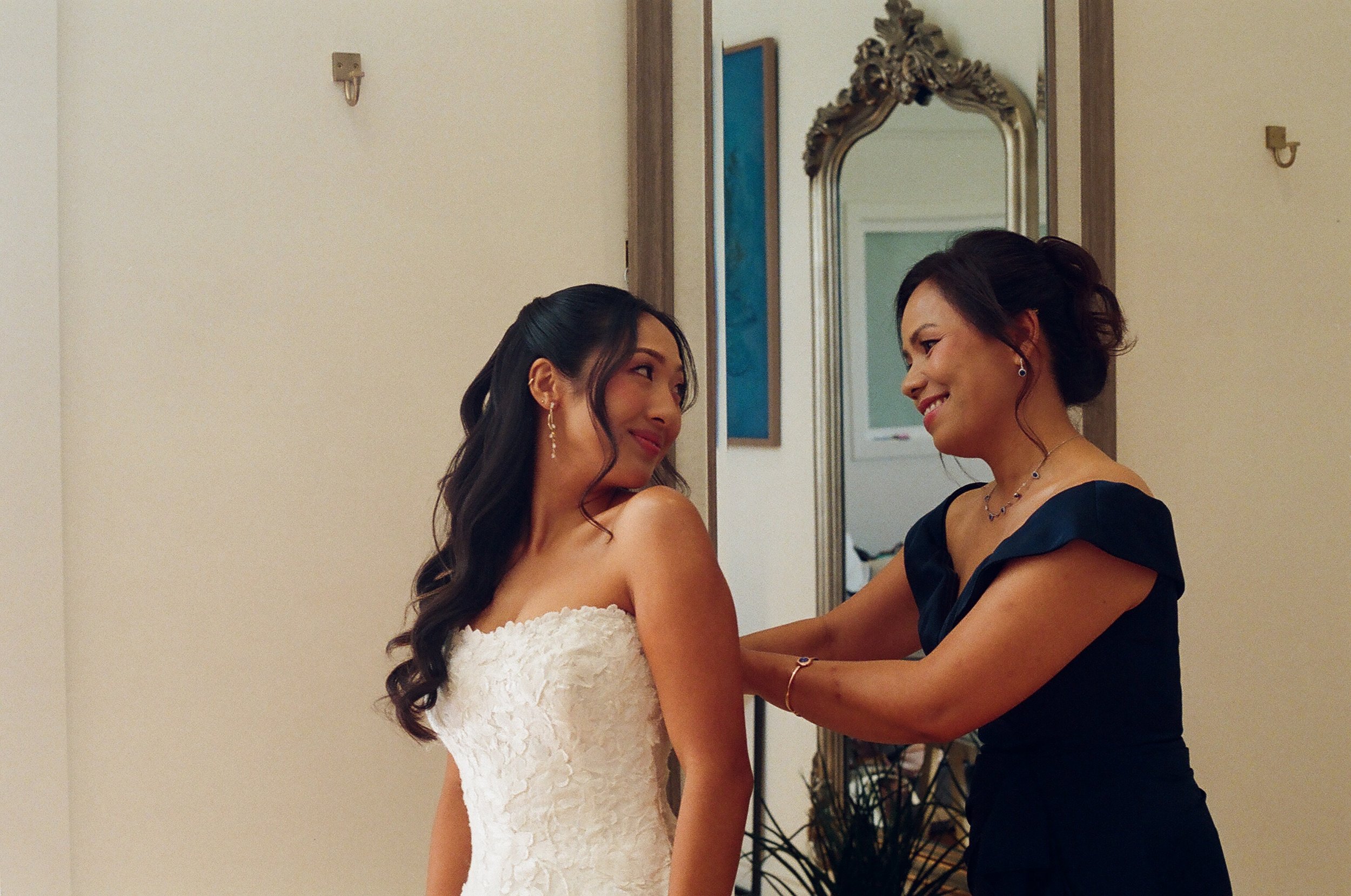 A bride in a white lace wedding dress with long, dark, curled hair and a woman in a black dress adjusting her dress inside a room with a mirror and wall art.