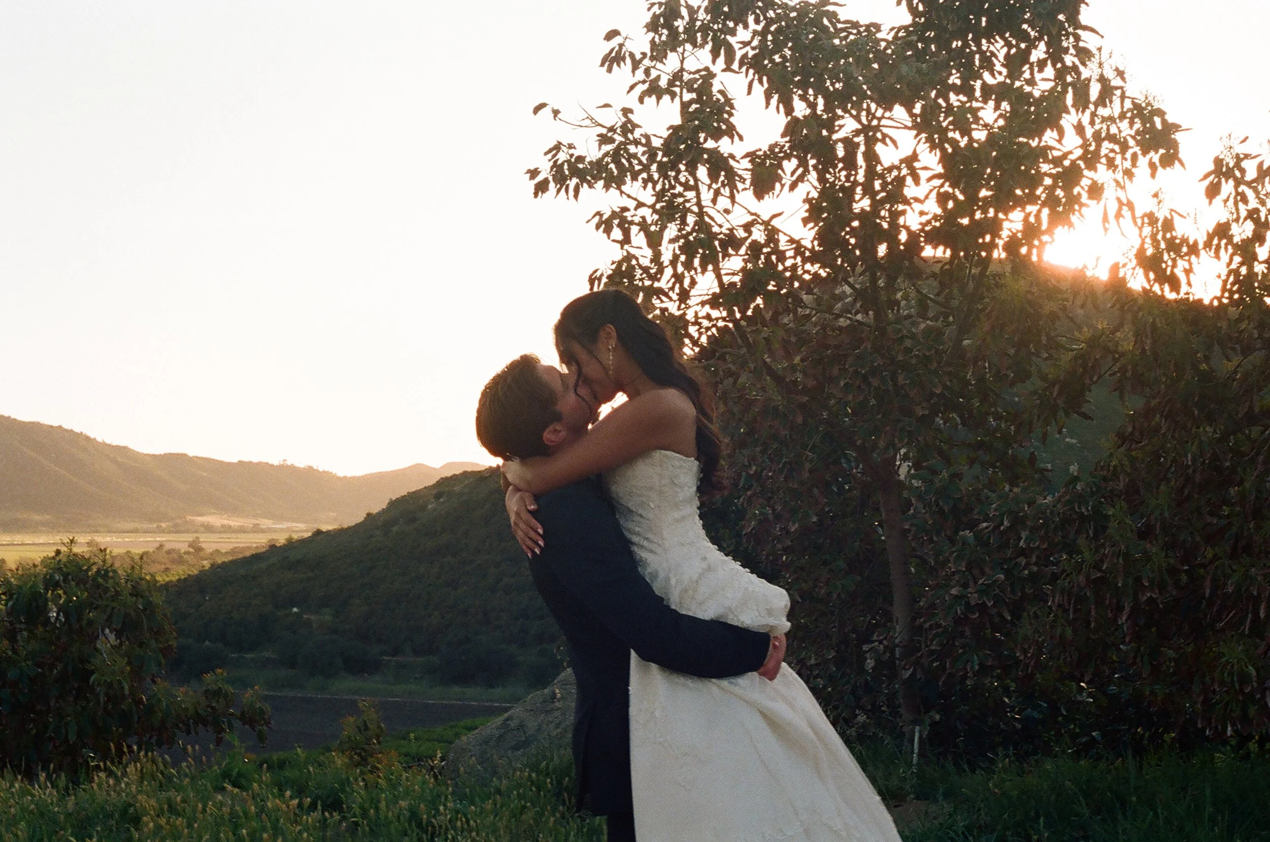 A couple dressed in wedding attire embracing and kissing outdoors during sunset, with trees, hills, and a clear sky in the background.