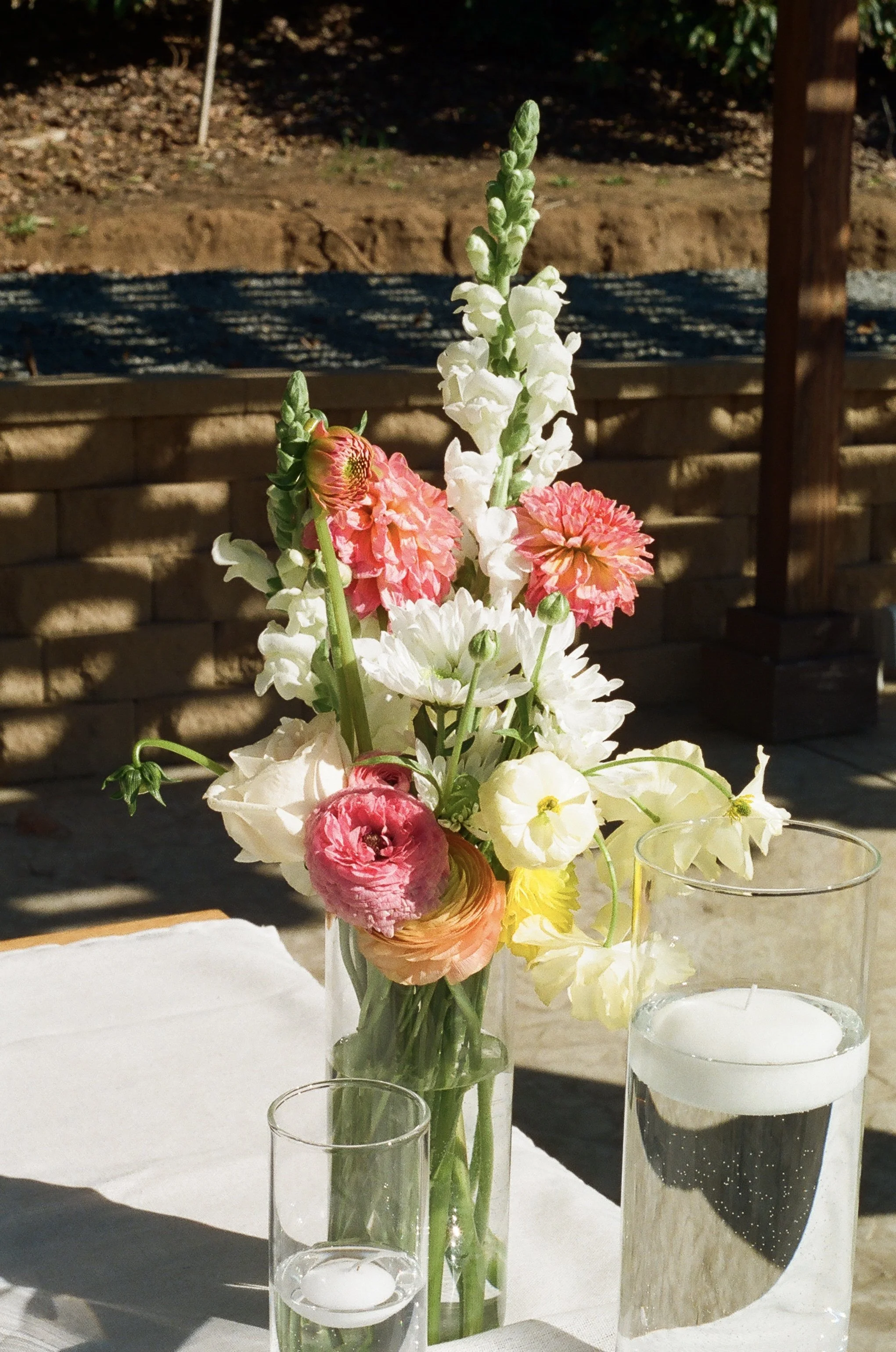 A floral centerpiece in a glass vase with white, pink, yellow, and peach blossoms on a table with candles in glass holders. The background shows a wooden fence and dirt ground outdoors.