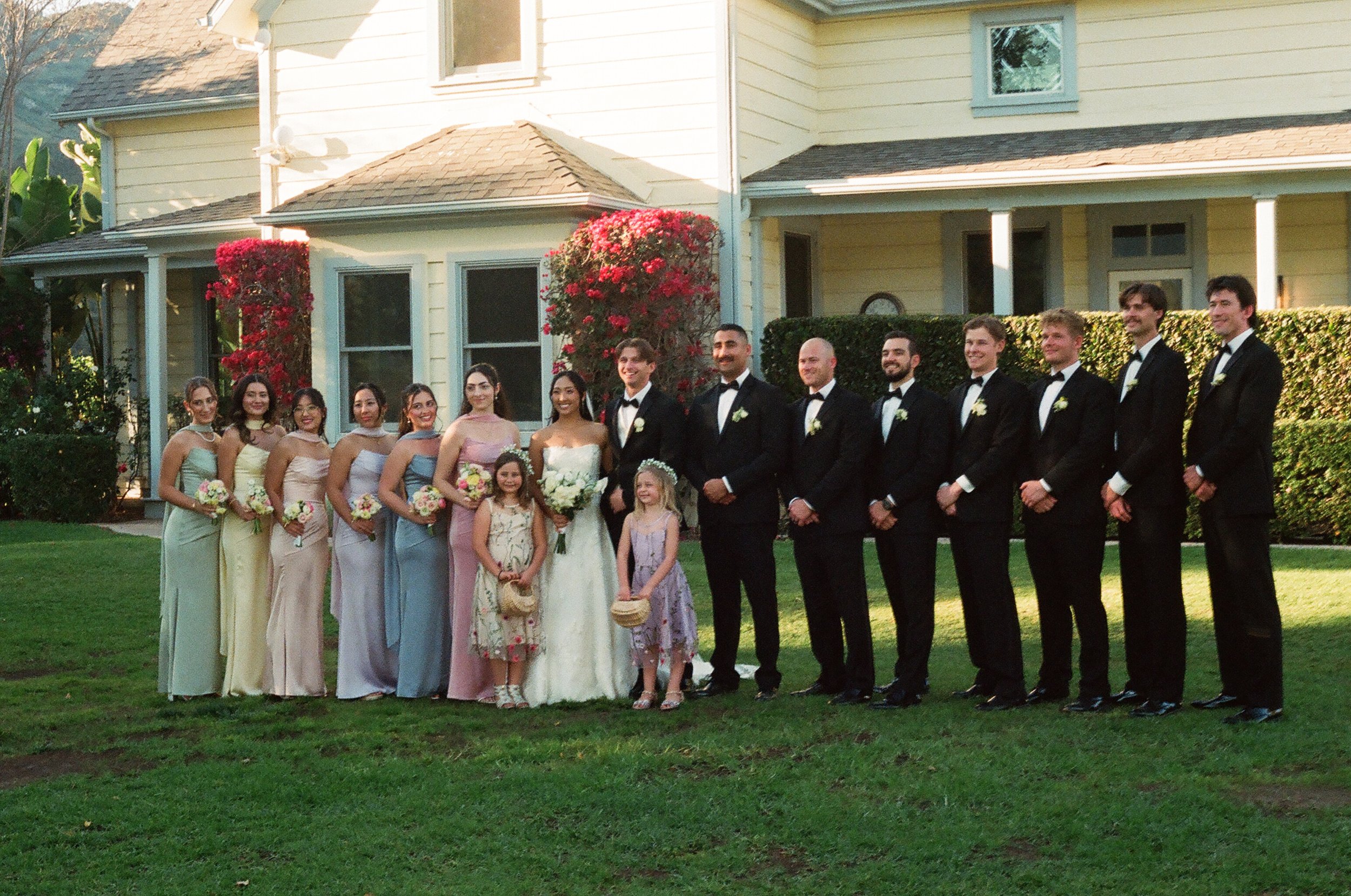 A group of young adults dressed for a wedding, standing outside in front of a yellow house with pink flowering bushes. The group includes women in colorful dresses, a bride in a white wedding gown, and men in tuxedos. The scene is during the daytime 