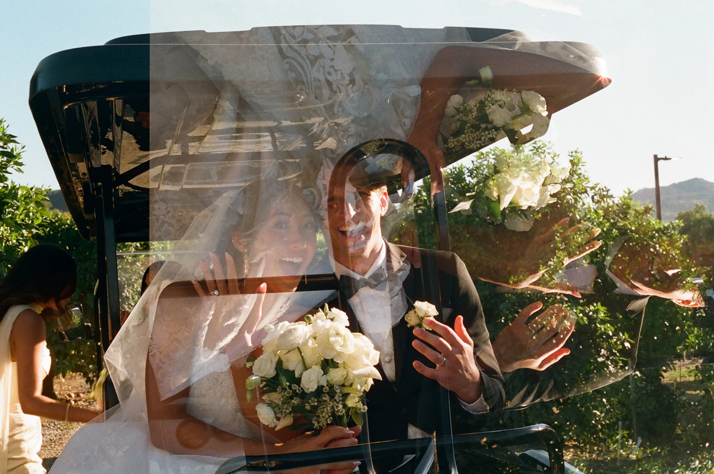 A newlywed bride and groom smiling and waving behind a glass hearse decorated with flowers, in an outdoor wedding setting surrounded by trees and sunlight.