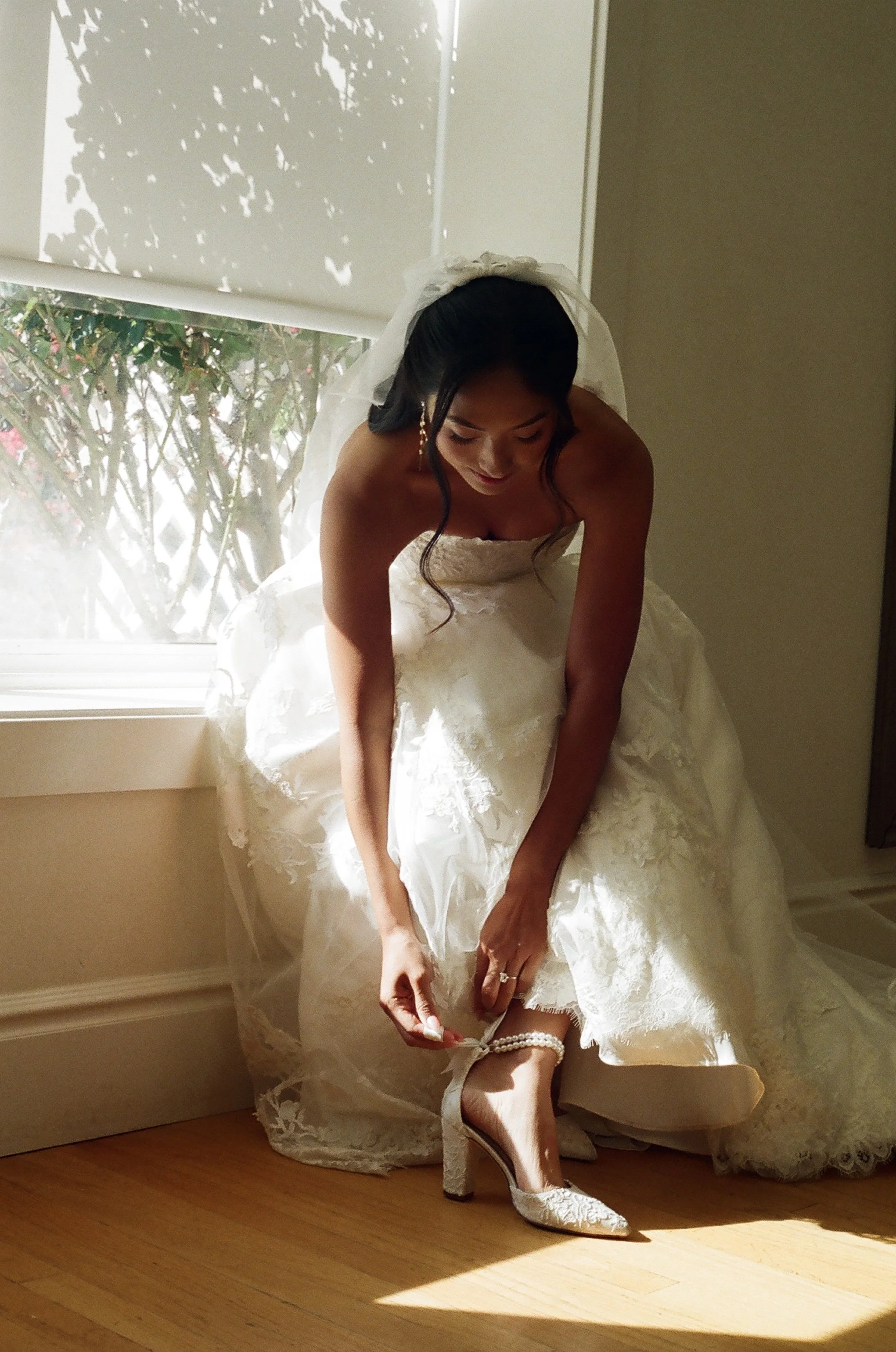 A bride in a white wedding dress is sitting on the floor by a window, adjusting her high-heeled shoe while holding a pearl bracelet. She has dark hair and is wearing earrings and a veil.
