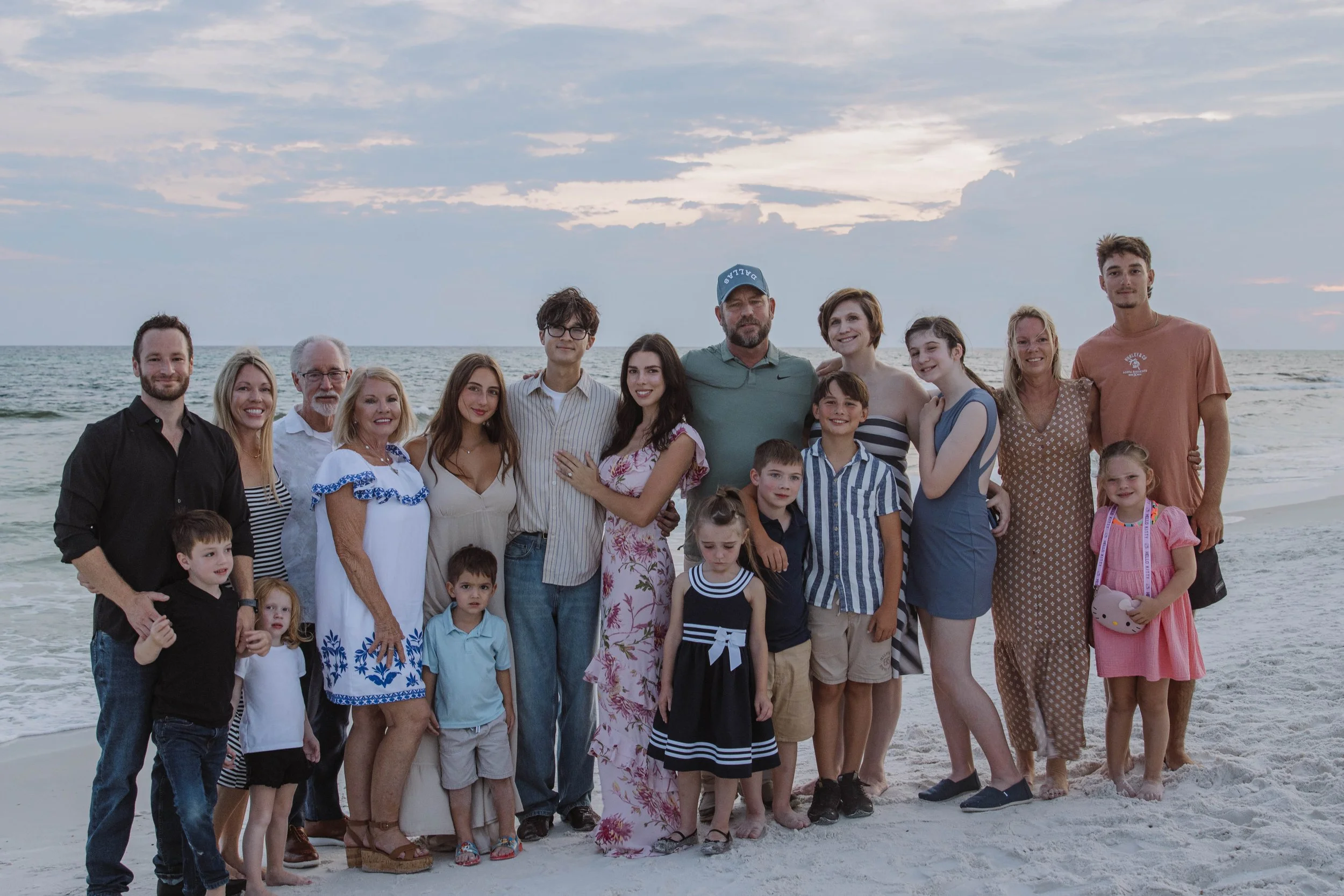 Group of people including children and adults standing on a beach at sunset with the ocean and partly cloudy sky in the background.