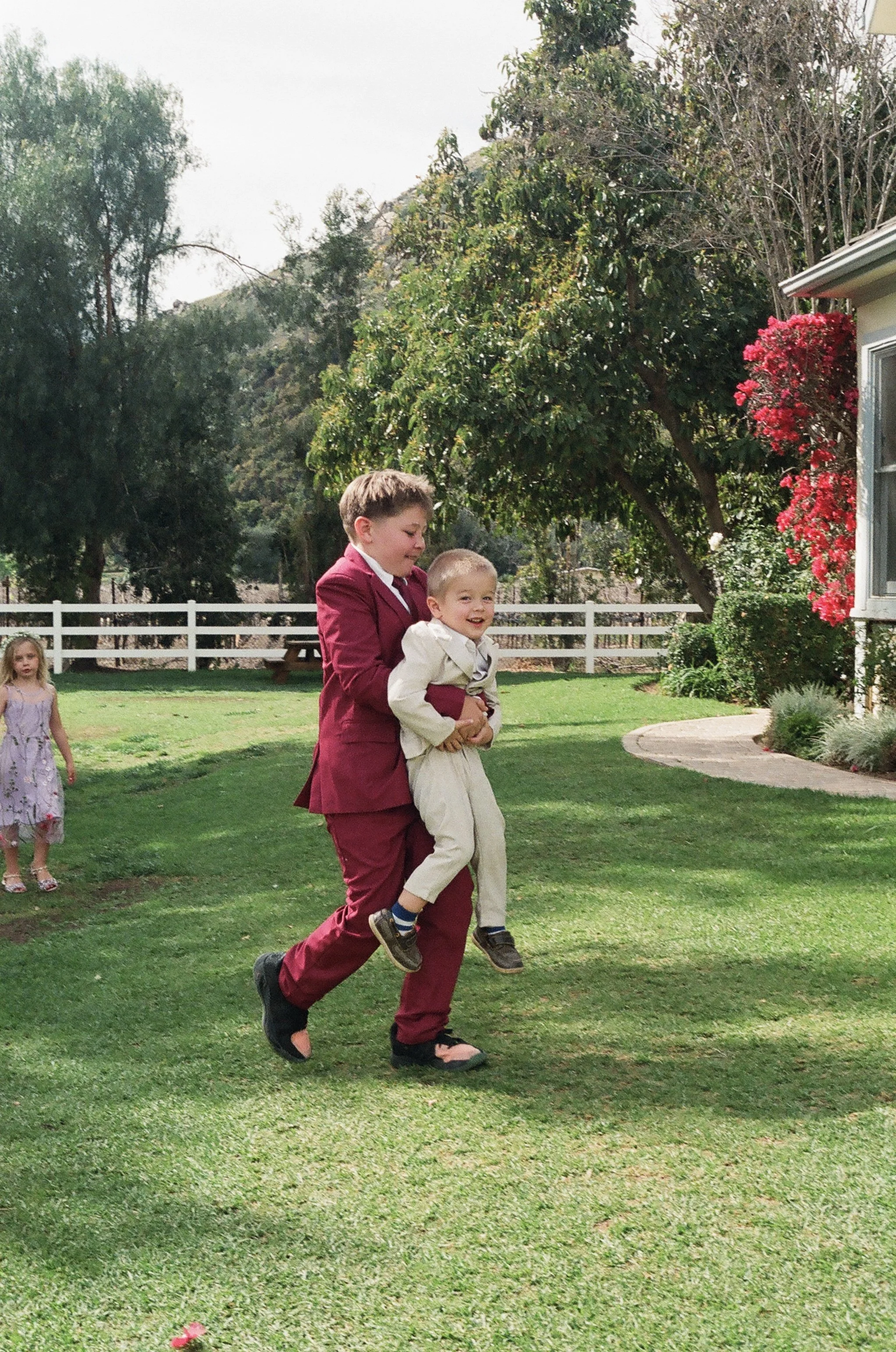 Two boys, one older in a maroon suit and the younger in a cream suit, playing outside on a grassy yard. The older boy is holding the younger boy in his arms as they walk or run. A young girl in a purple dress stands in the background near a white fen