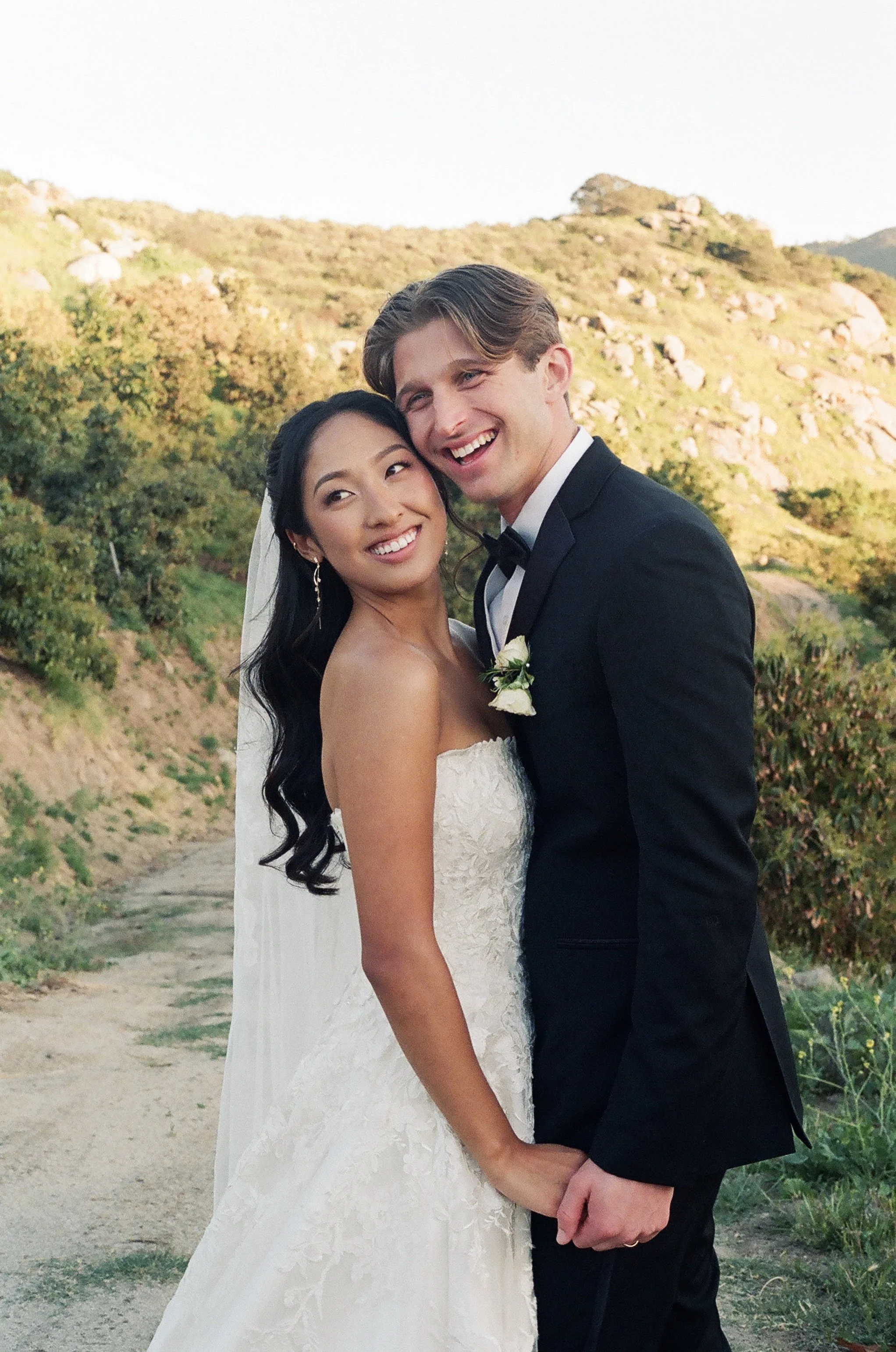 A smiling bride and groom holding hands outdoors with green hills and rocks in the background at sunset.