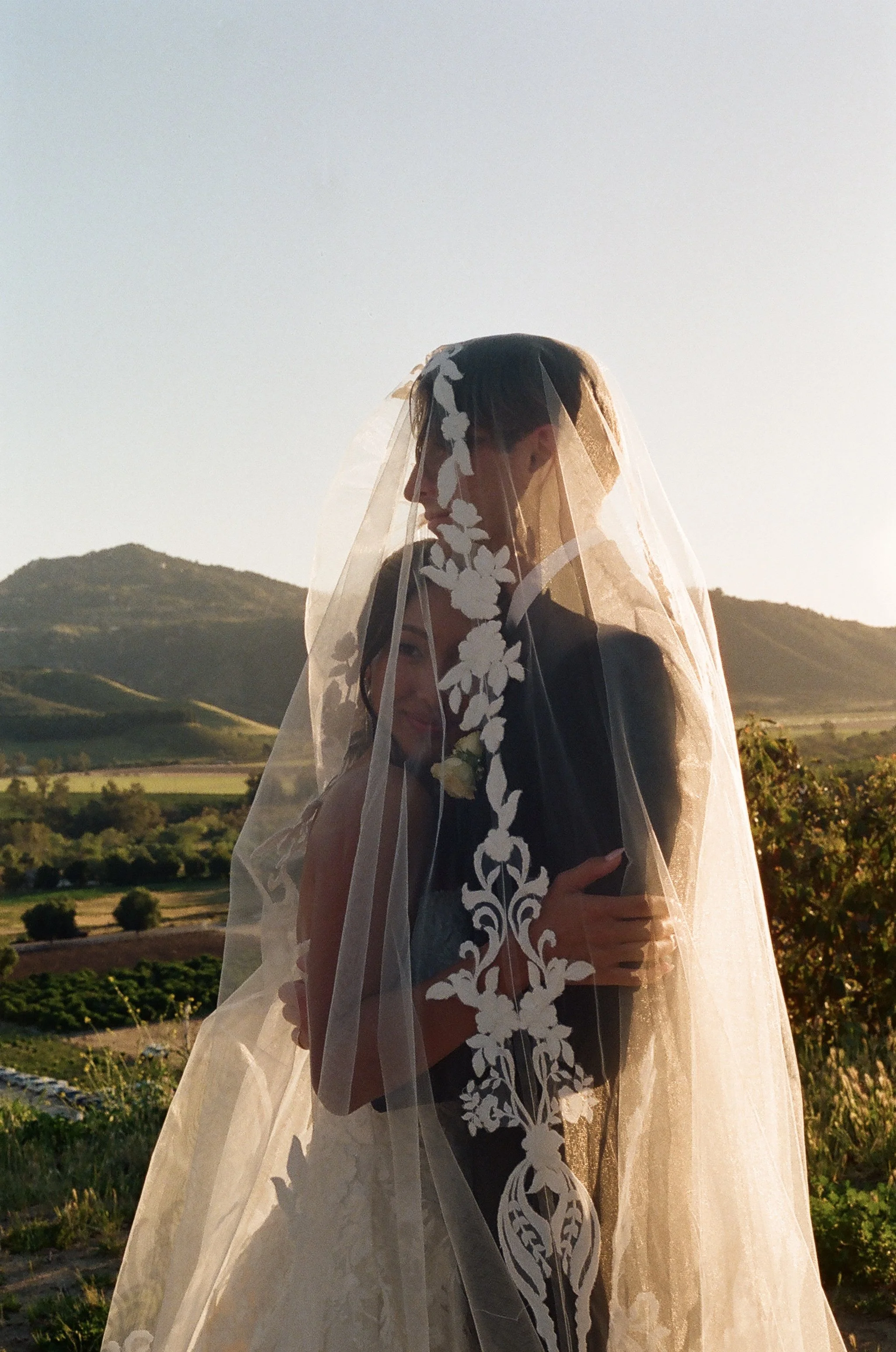 A woman in a wedding dress with a lace veil standing outdoors during sunset, with rolling hills and farmland in the background.