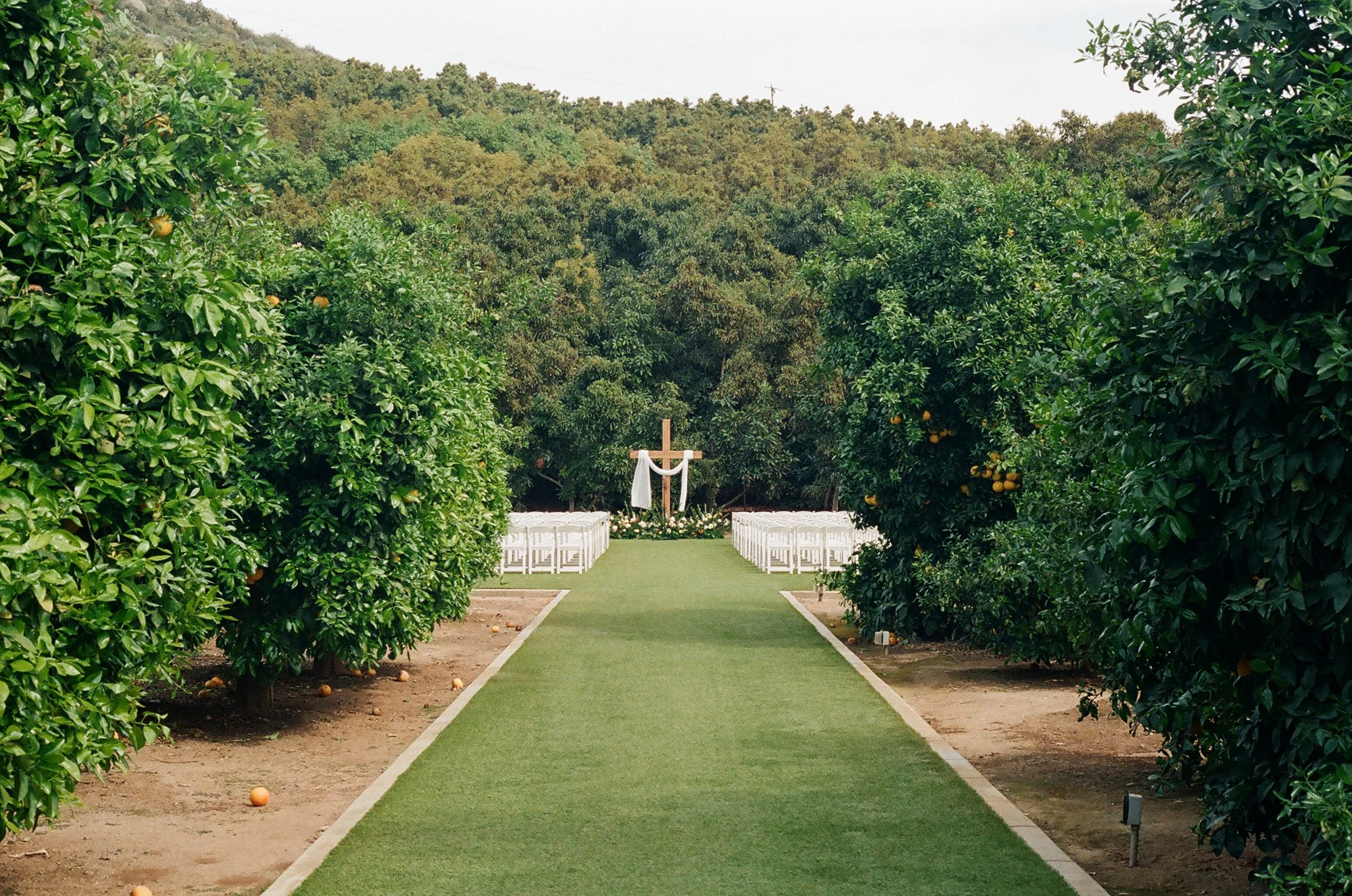 A miniature golf course with an artificial grass path, trees on both sides, and white chairs arranged in rows for seating, with a cross and cloth in the background.