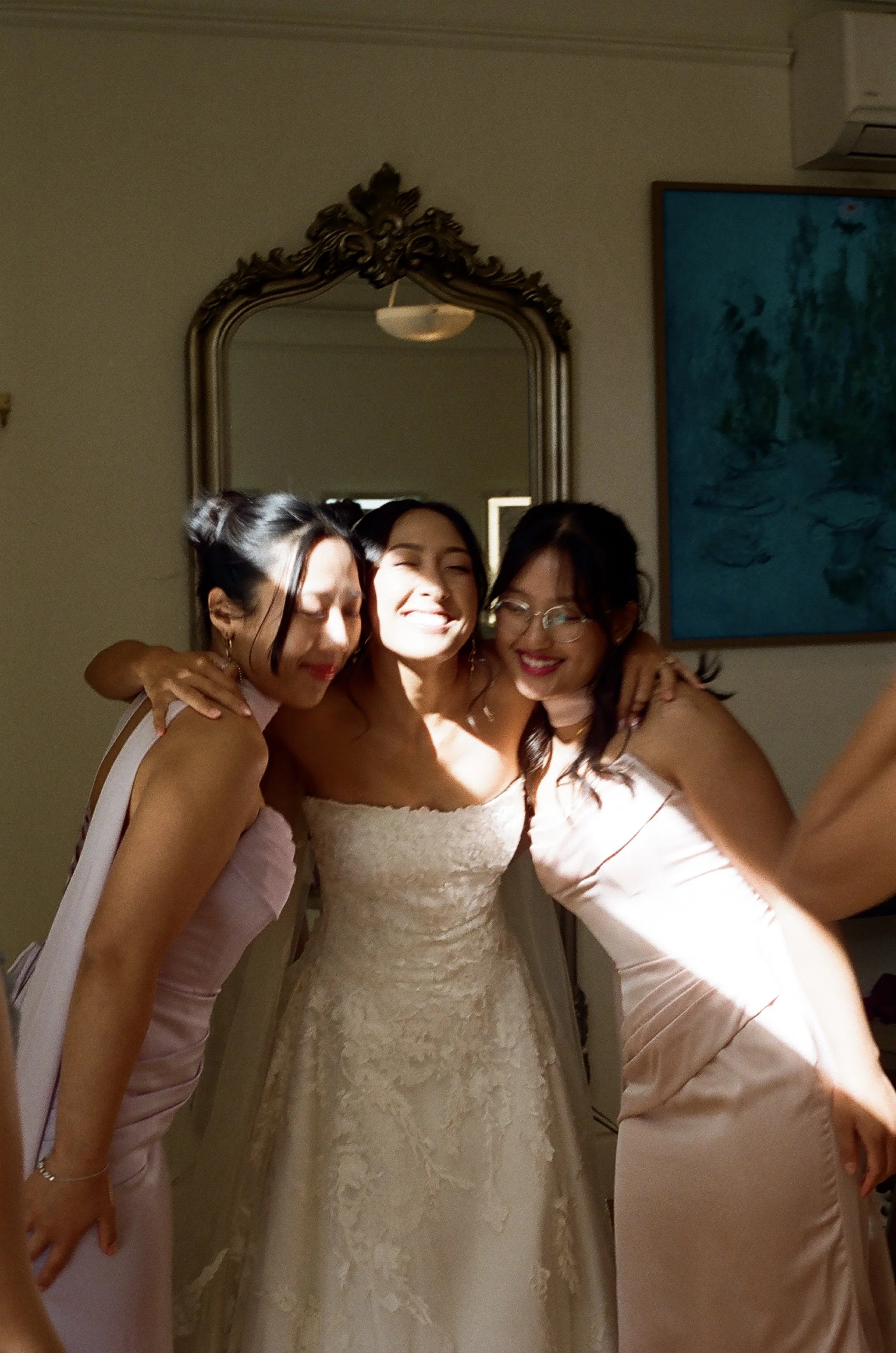 Three women, one in a white wedding dress, celebrating and hugging each other indoors.