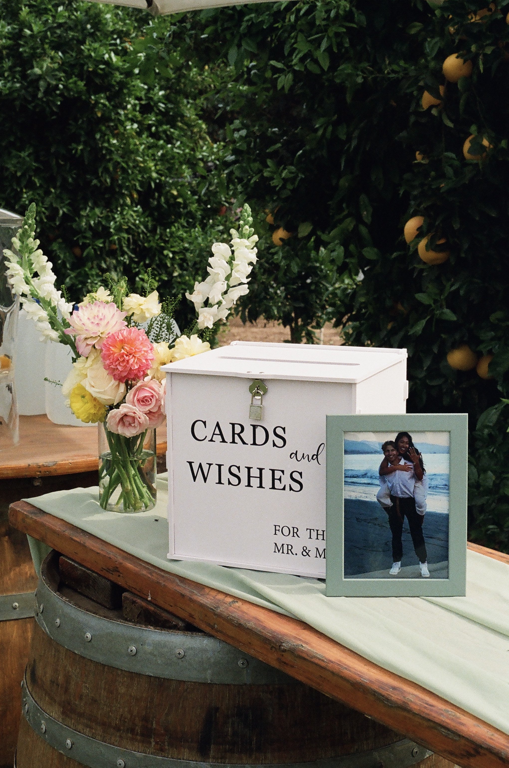 A bouquet of pink and yellow flowers in a clear vase on a wooden barrel, with a wedding card and a framed photo of a couple at the beach next to it.