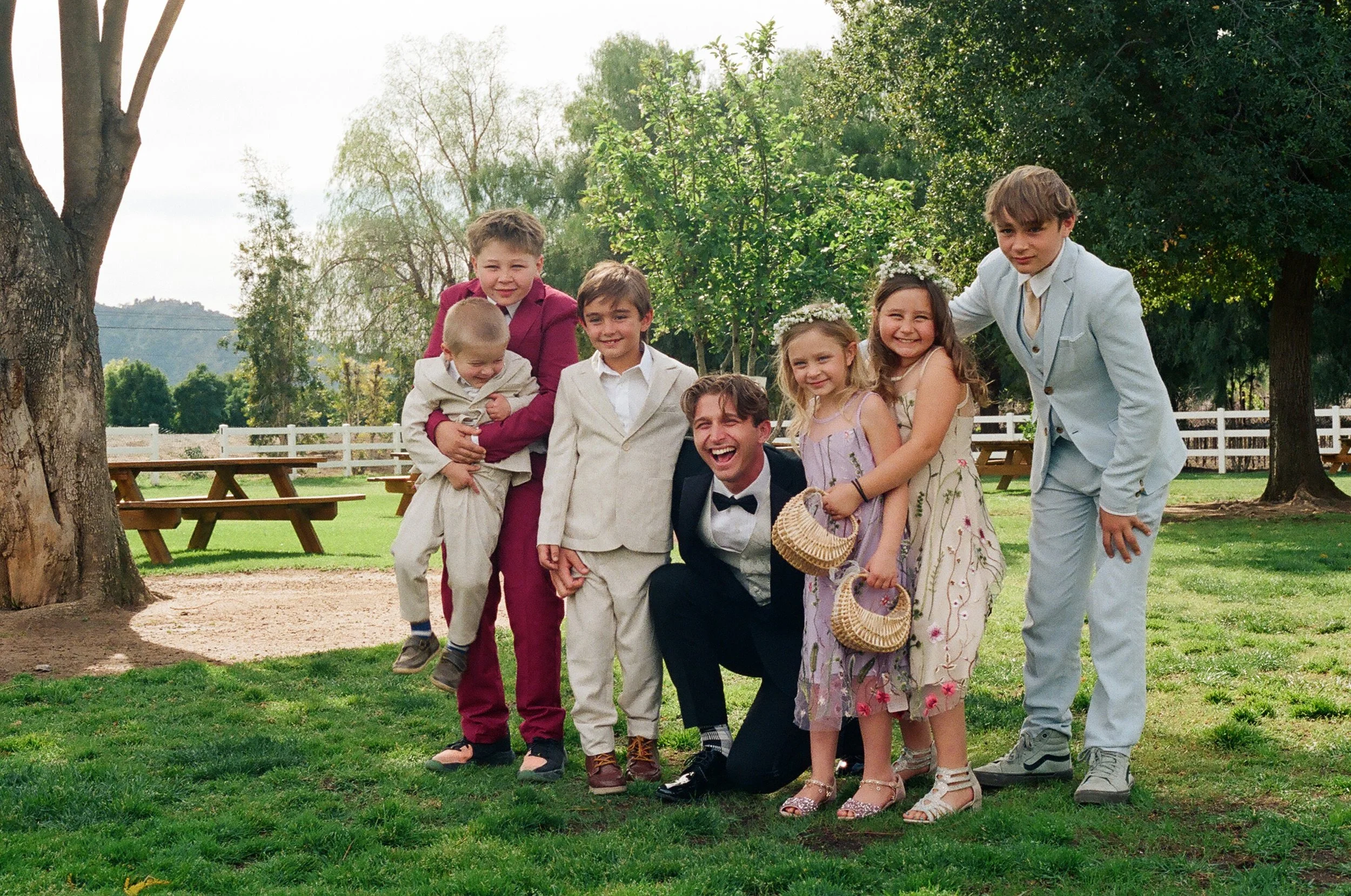 A group of children and a man, dressed in semi-formal and formal clothing, pose outdoors on a grassy area with trees and picnic tables in the background. The children are smiling and holding small baskets, suggesting a special occasion or celebration