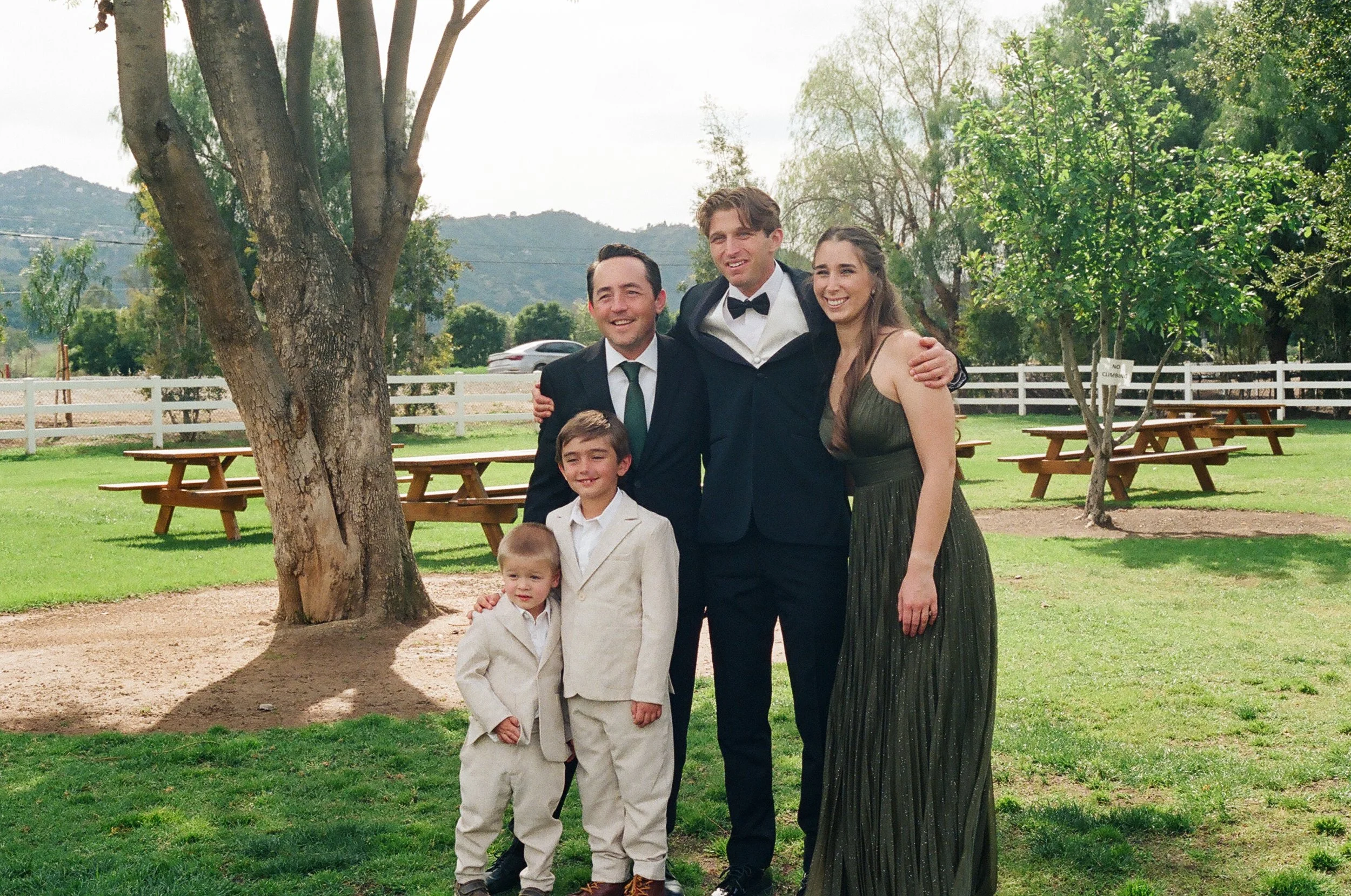 A group of five people, including three adults and two children, standing outdoors under a large tree during daytime. They are dressed in formal attire, with the adults in suits and the children in light-colored suits, smiling and posing for a photo 