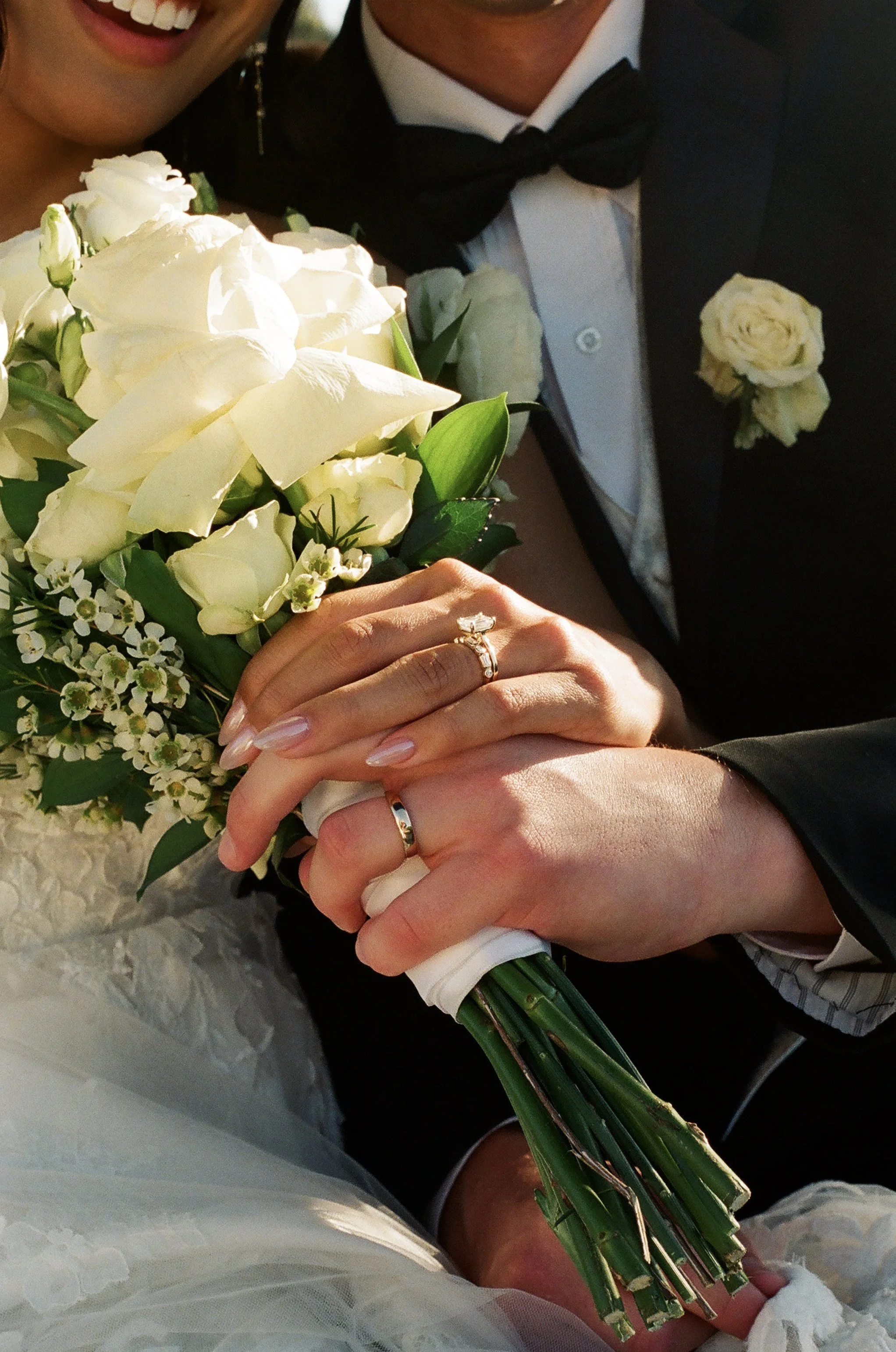 Close-up of a bride and groom holding a bouquet of white flowers with their hands intertwined, showing wedding rings, in a joyful moment.