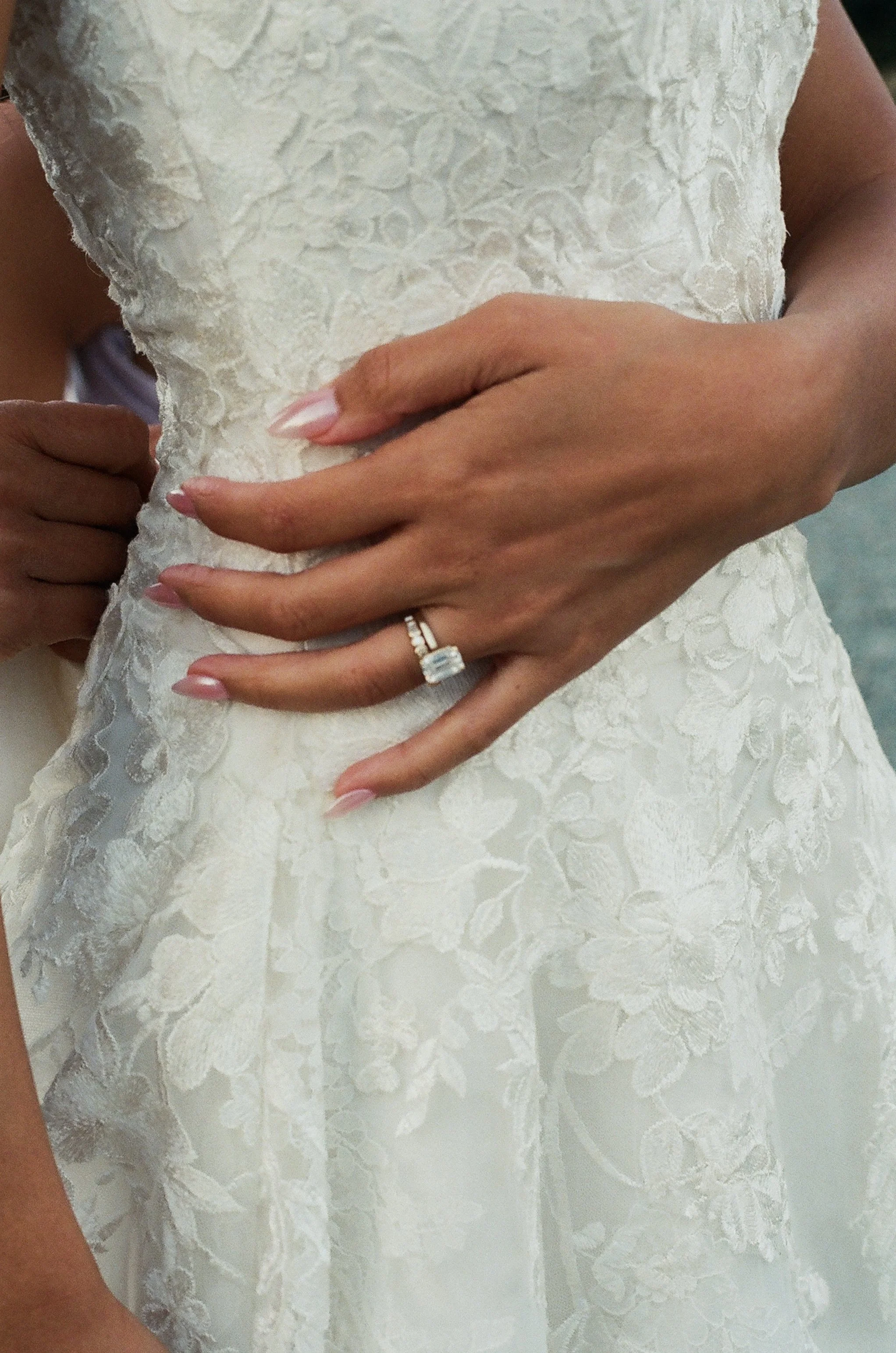 Close-up of a bride wearing a white lace wedding dress, with hands resting on her waist, one hand showing a wedding ring and manicured nails.