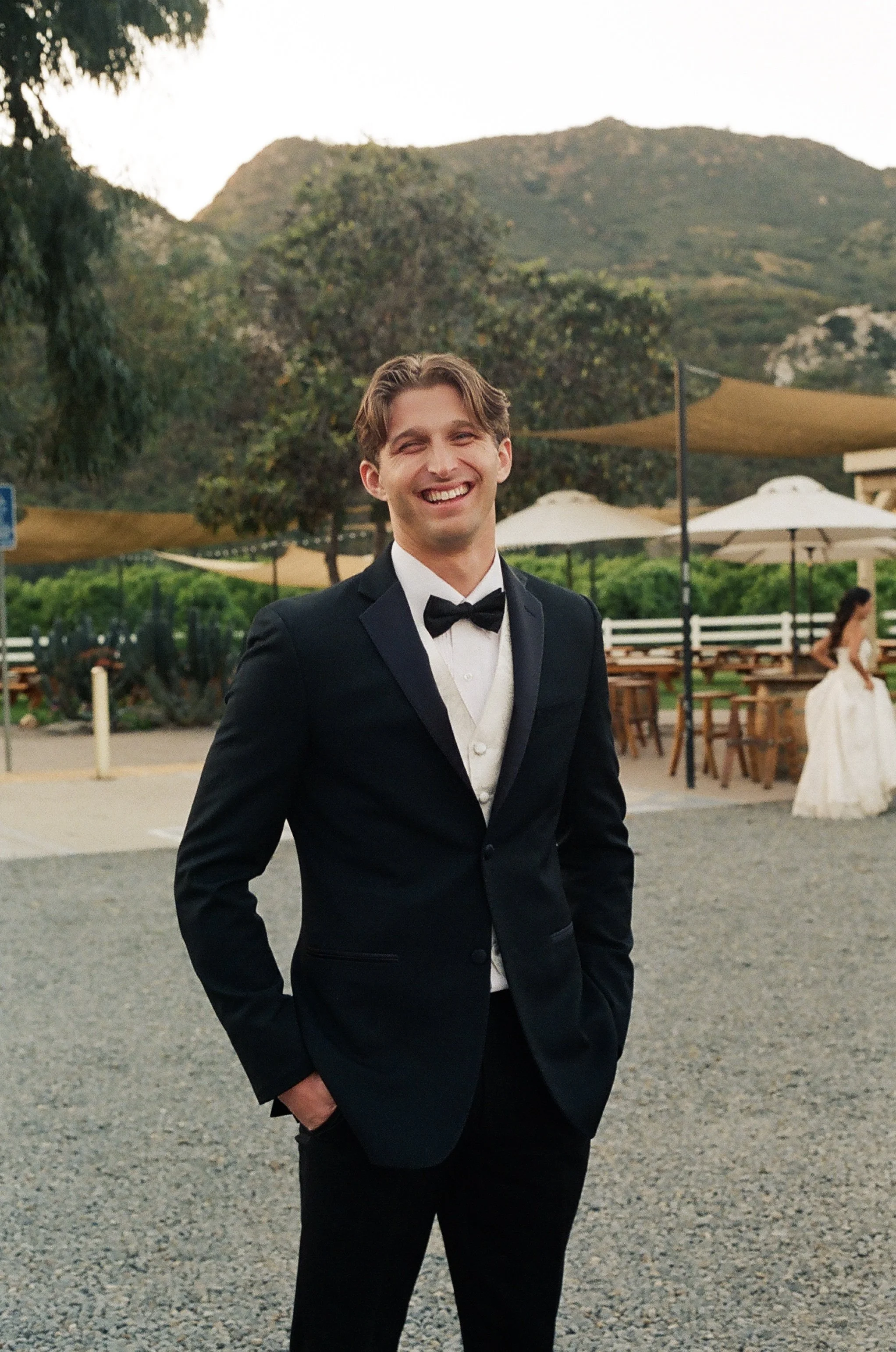 A young man in a black tuxedo with a bow tie standing outdoors in front of trees and mountains, smiling.