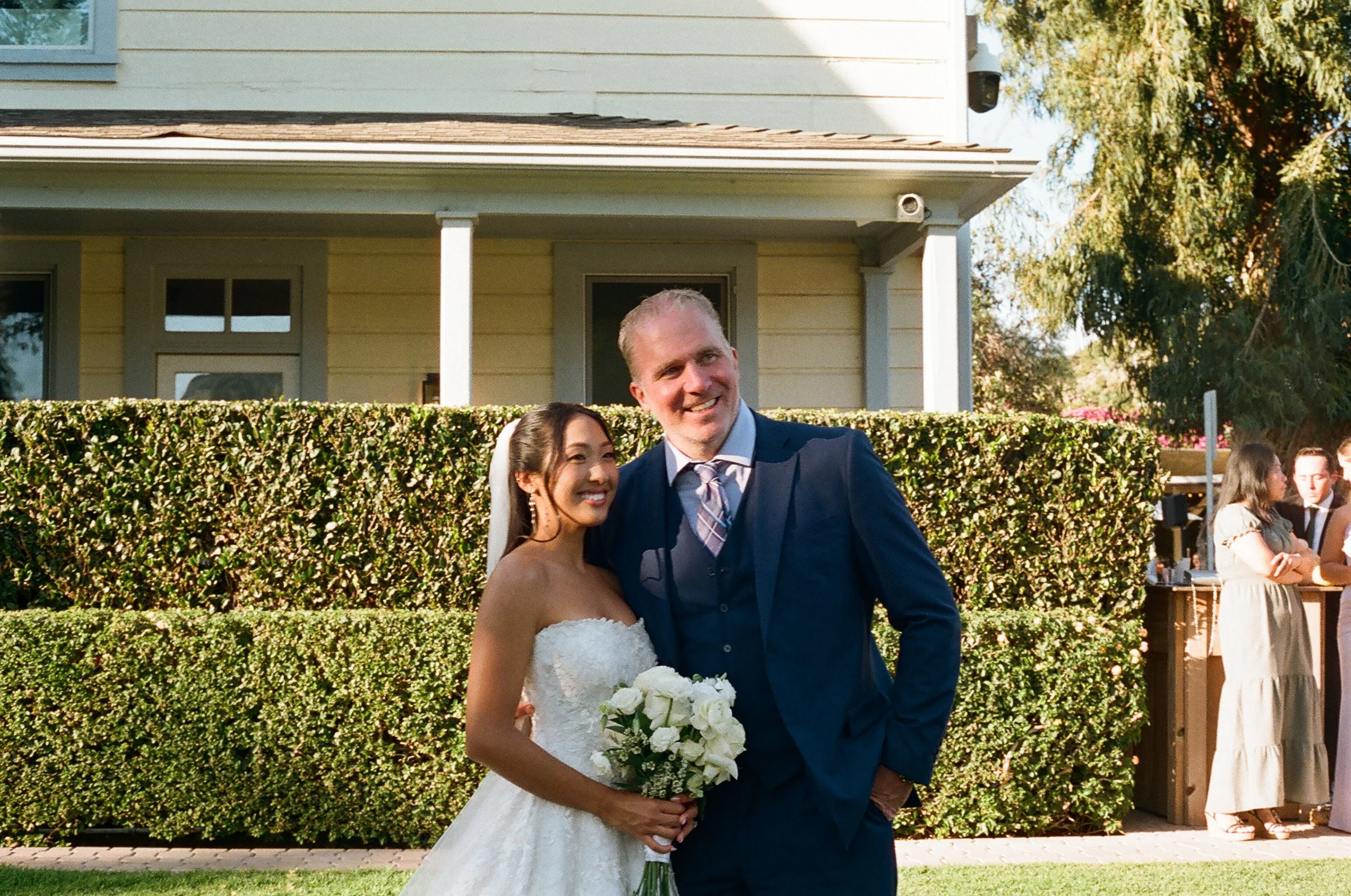 A smiling bride and groom posing outdoors at a wedding, with a house and other wedding guests in the background.