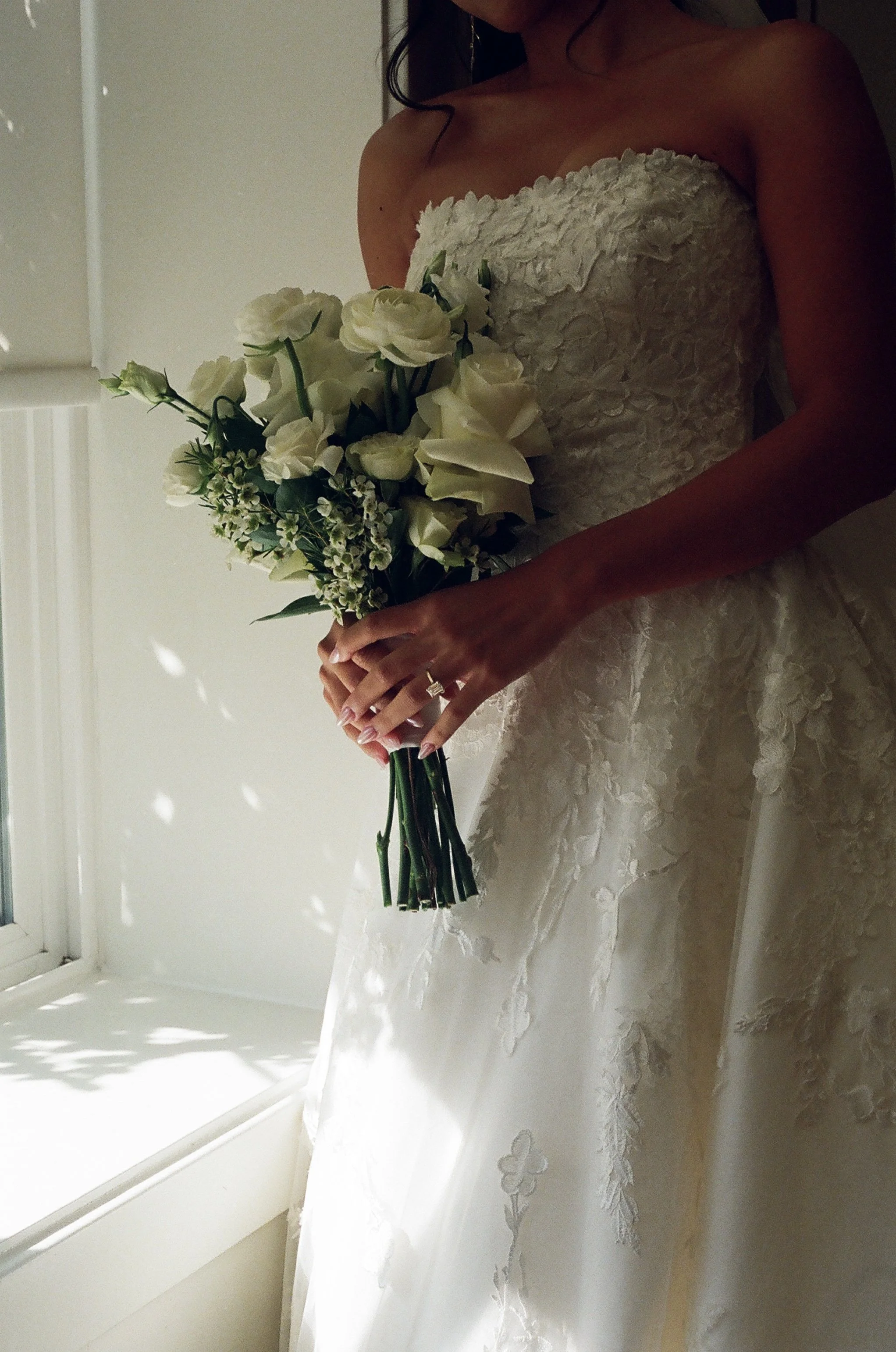 A woman in a white wedding dress holding a bouquet of white flowers, standing near a window with sunlight casting shadows.