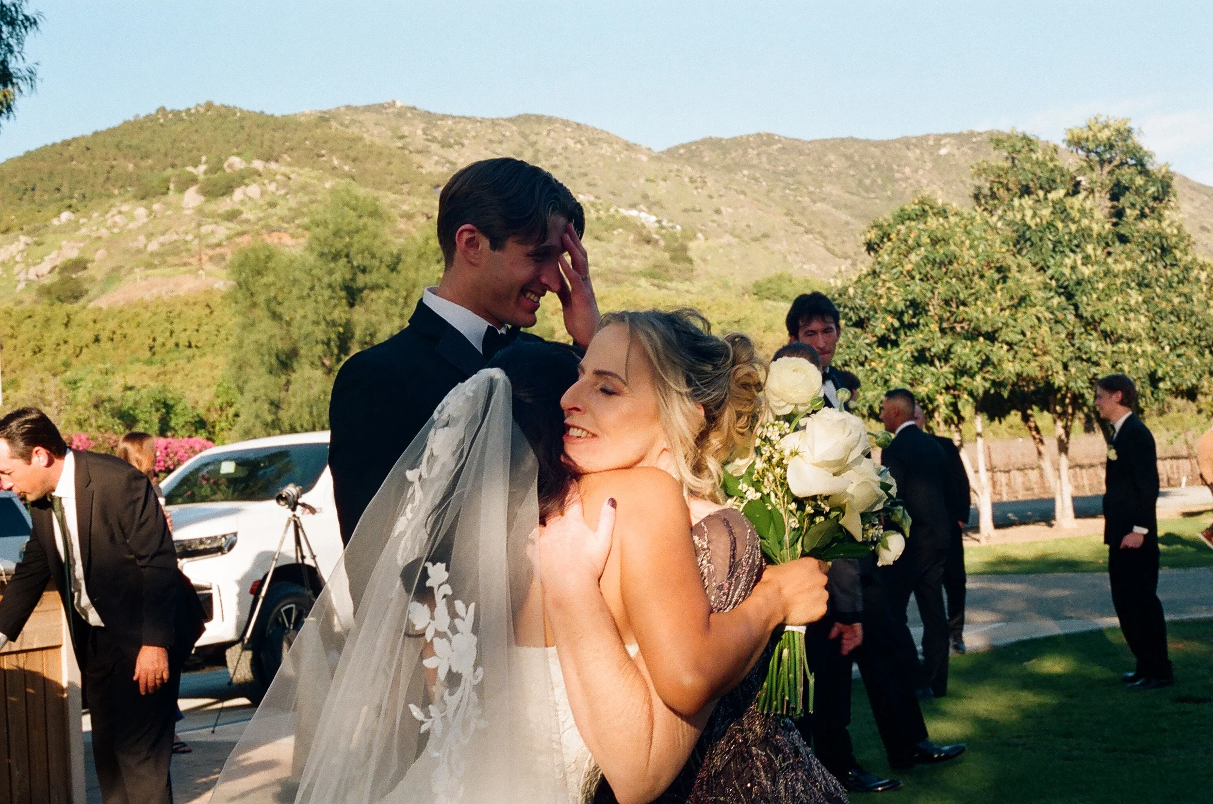 A bride and groom sharing a hug outdoors during their wedding celebration, with potential mountains and guests in the background.