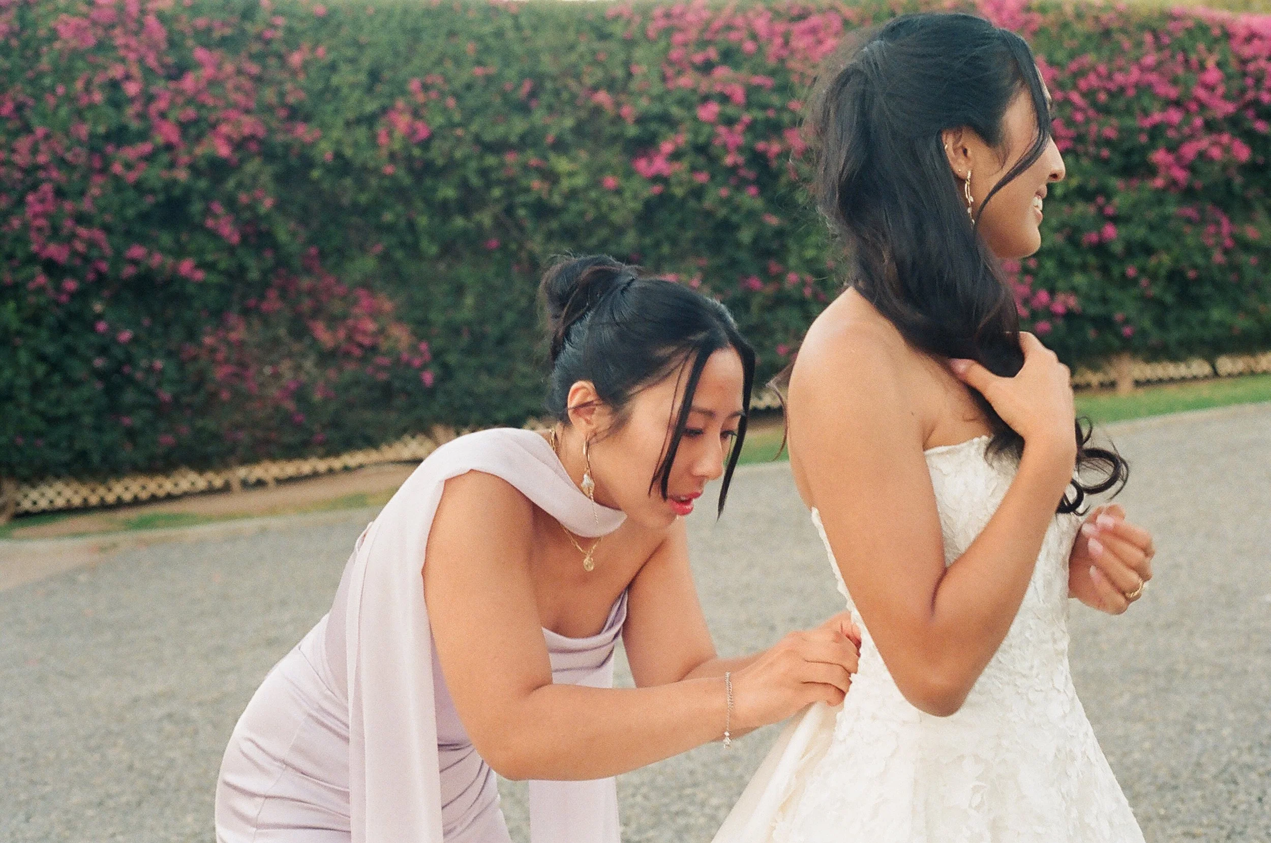 Two women dressed in white, kneeling outdoors, one of them in a wedding dress, with a background of pink flowering bushes.