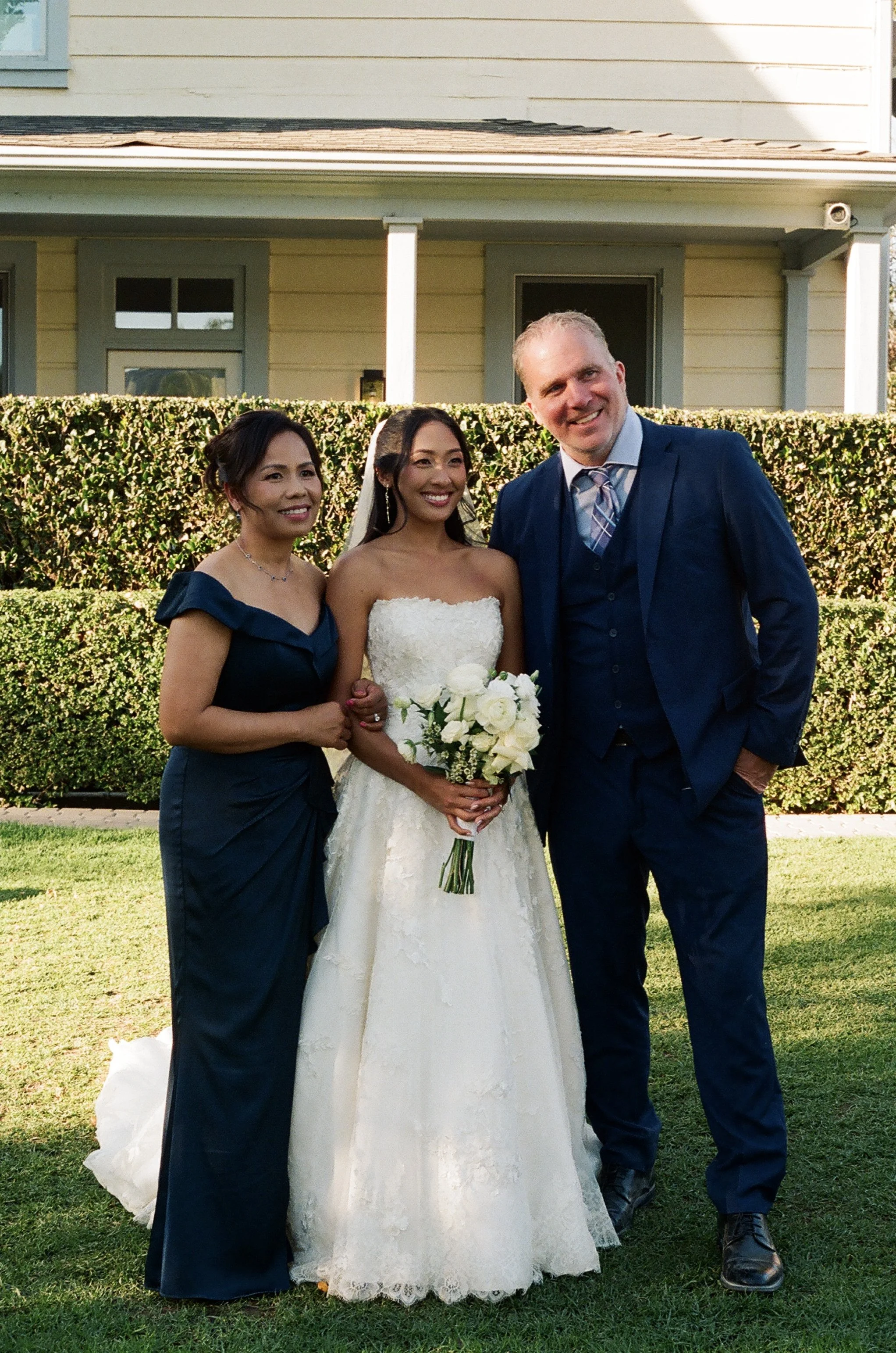 A bride in a lace strapless wedding dress holding a bouquet of white roses, standing between a woman in a navy dress and a man in a navy suit, in front of a hedge and house.