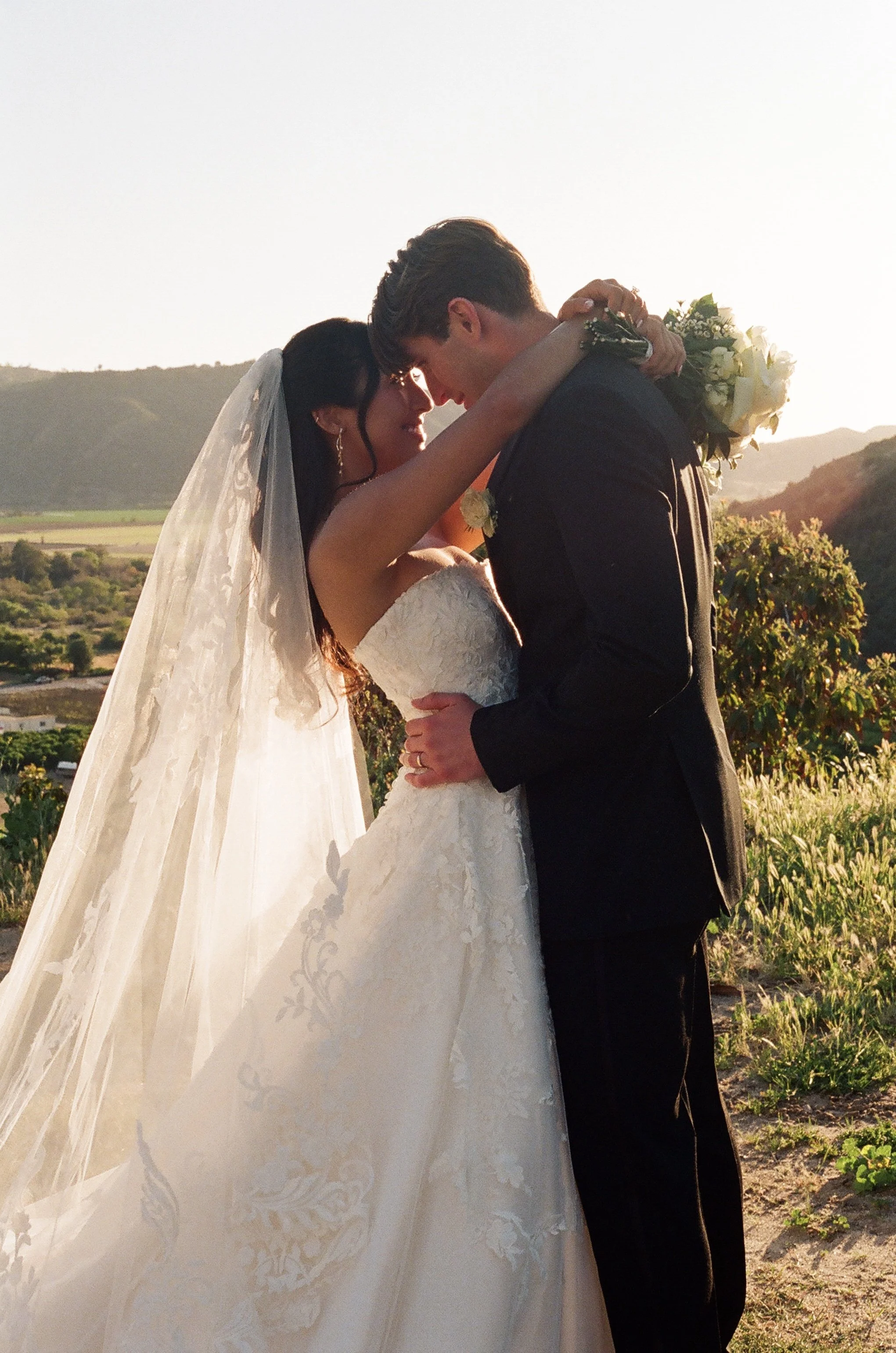 A bride and groom sharing a kiss outdoors at sunset, with mountains and greenery in the background.