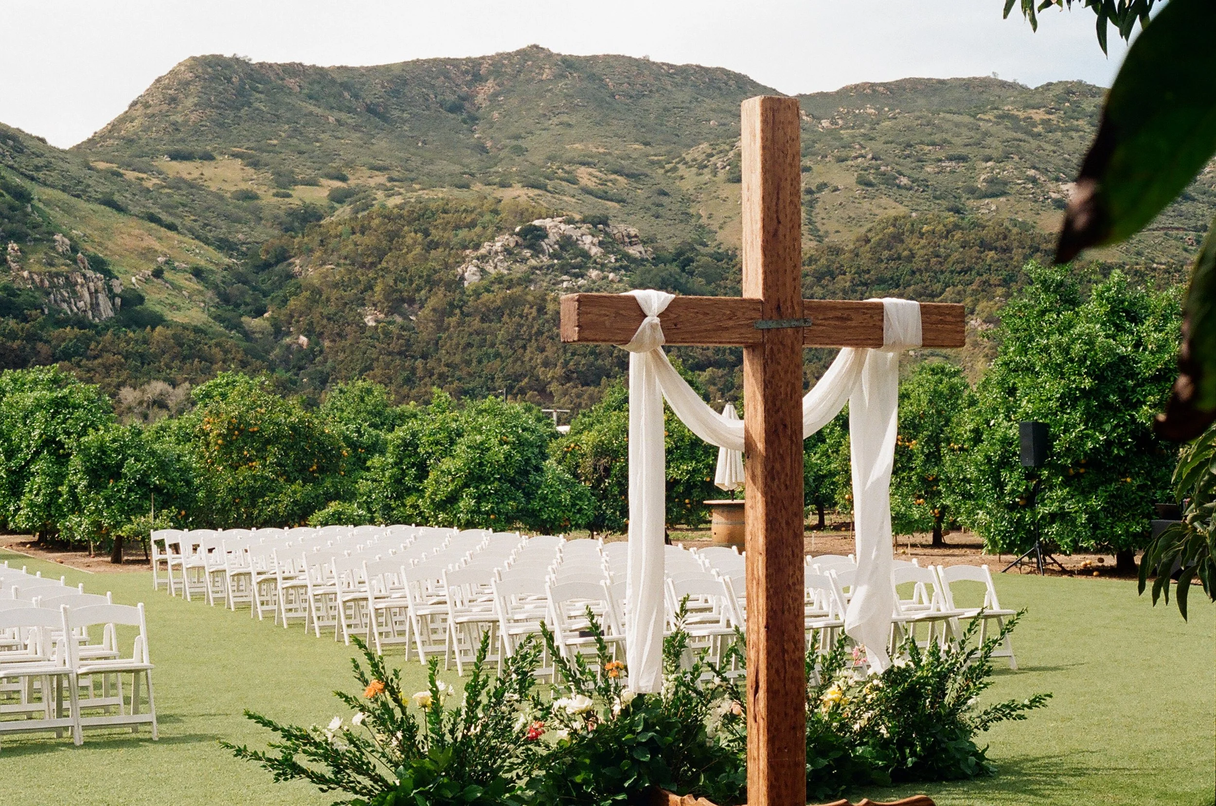 A wooden wedding cross is decorated with white fabric ribbons and is set outdoors amidst rows of white chairs, with mountains and greenery in the background.
