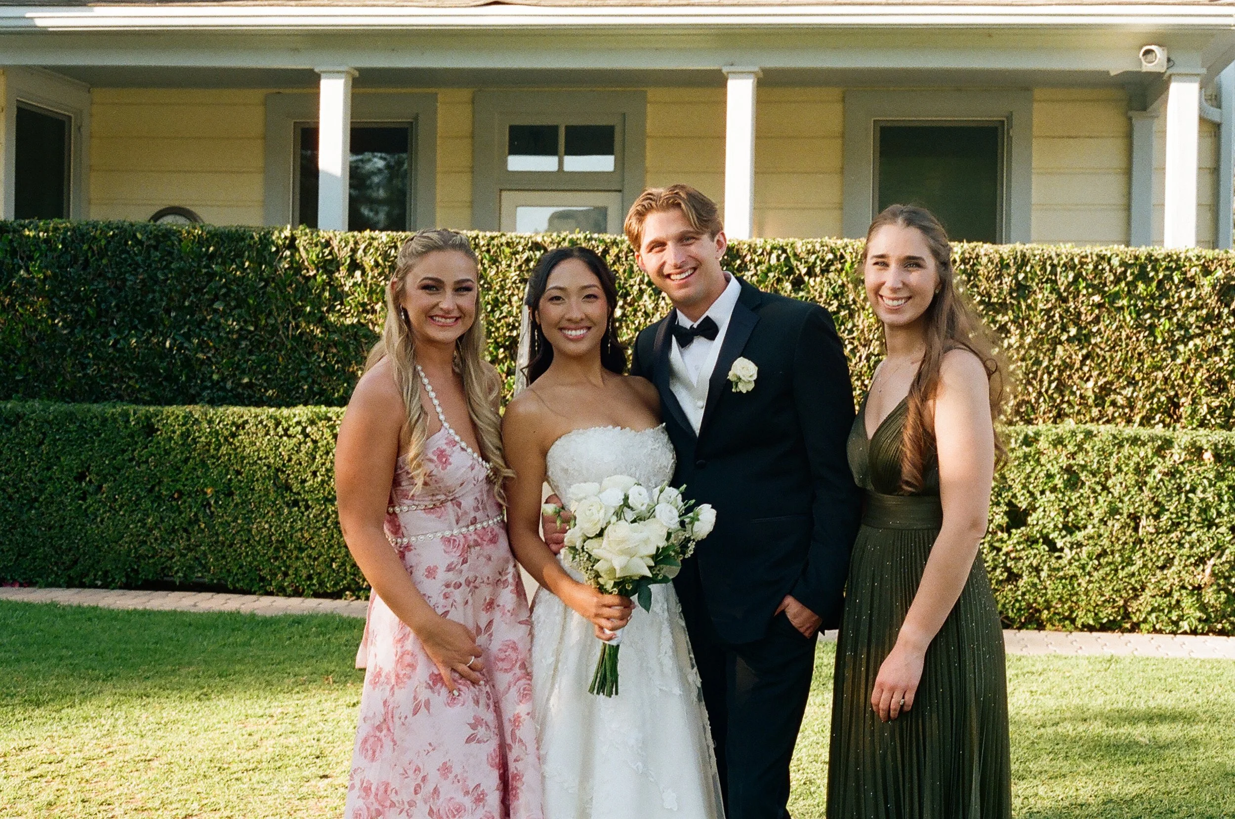 A group of four people, including a woman in a white wedding dress holding a bouquet of white flowers, standing outdoors in front of a hedge and a light-colored house with a front porch. The group appears to be celebrating a wedding.