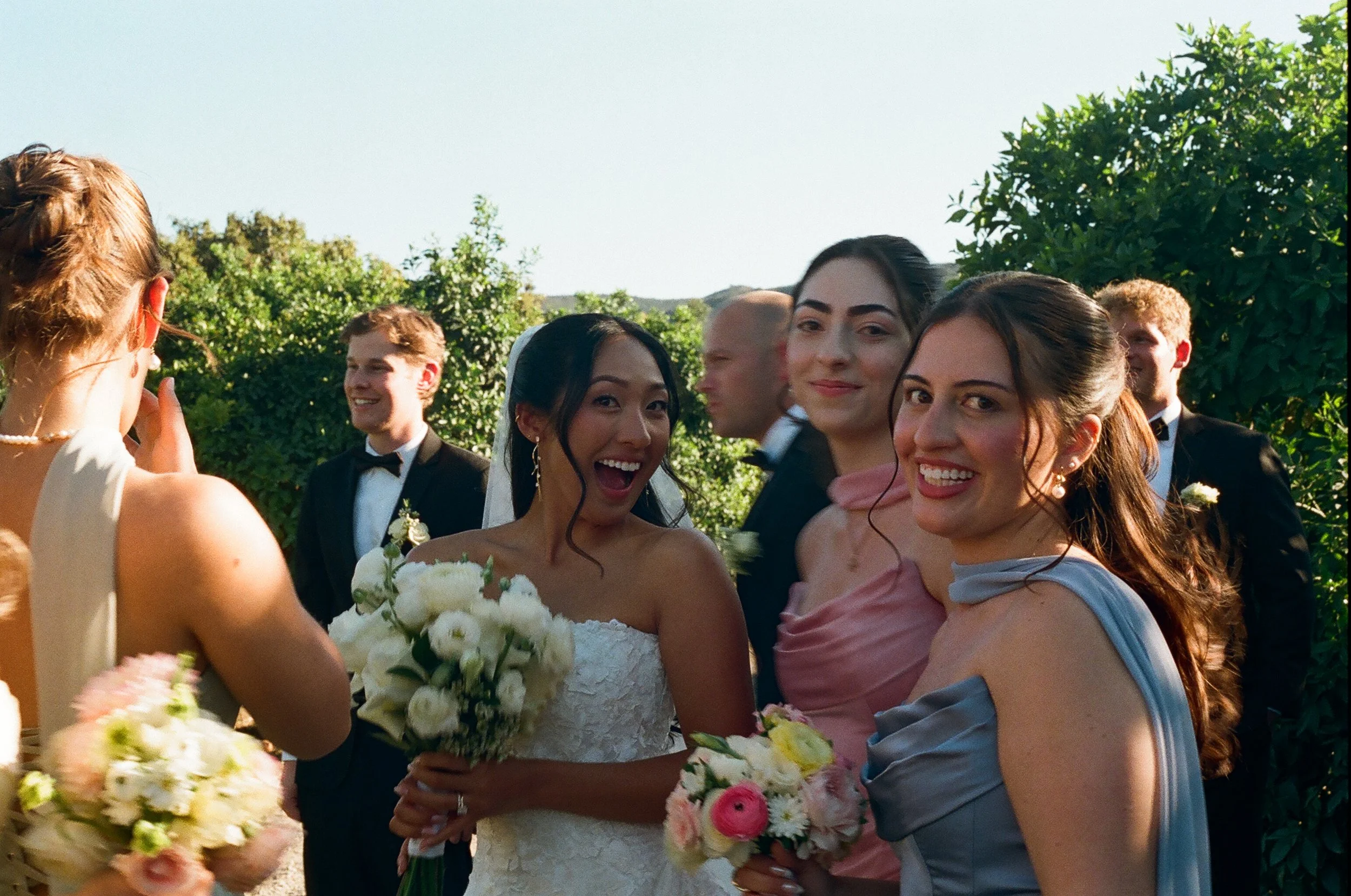 Group of people at a wedding, including a bride in a white dress holding a bouquet, and women in pastel-colored dresses, smiling and interacting outdoors with green trees in the background.