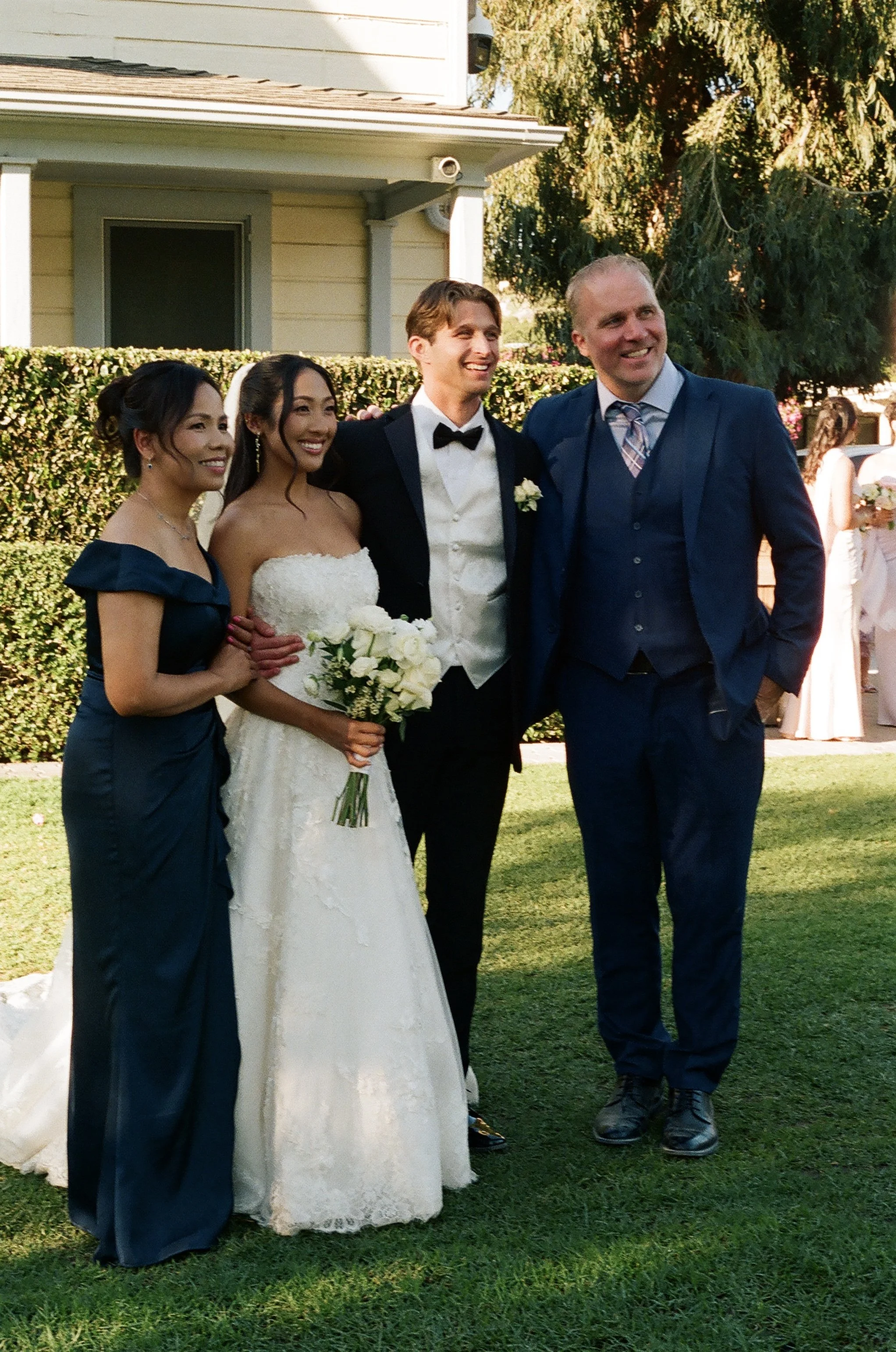 A group of four people standing outside on a grassy area during a wedding. There is a bride in a white strapless wedding gown holding a white bouquet and two women in matching dark blue dresses. Two men are dressed in suits, one in a tuxedo and the o