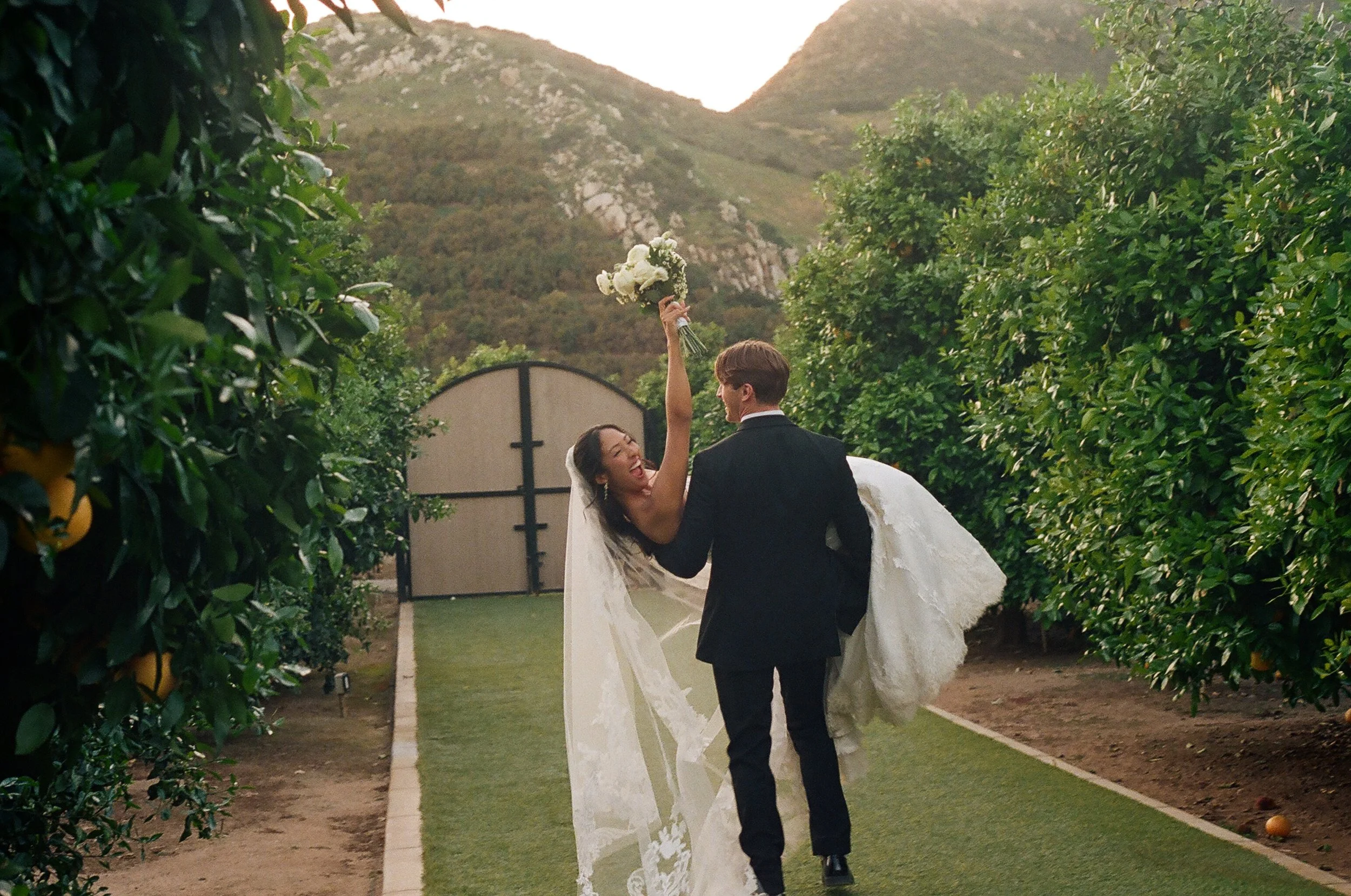 A bride and groom standing outdoors on a wedding day, with the bride holding a bouquet and the groom lifting her, surrounded by lush greenery and mountains in the background.