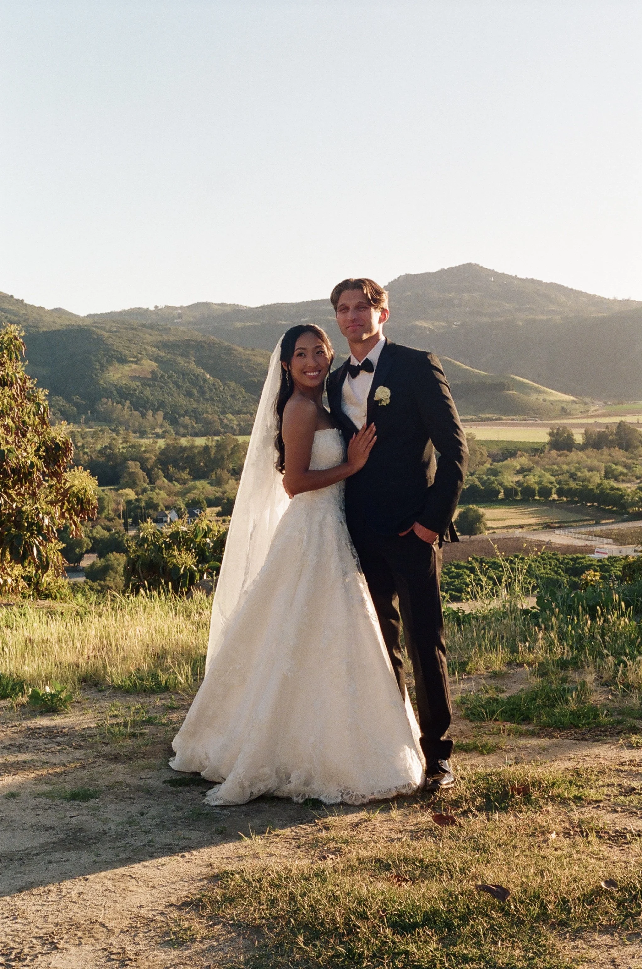A bride and groom standing outdoors on a grassy area with mountains in the background, during sunset. The bride is wearing a white strapless wedding dress and veil, smiling, and the groom is dressed in a black tuxedo with a bow tie, standing beside h