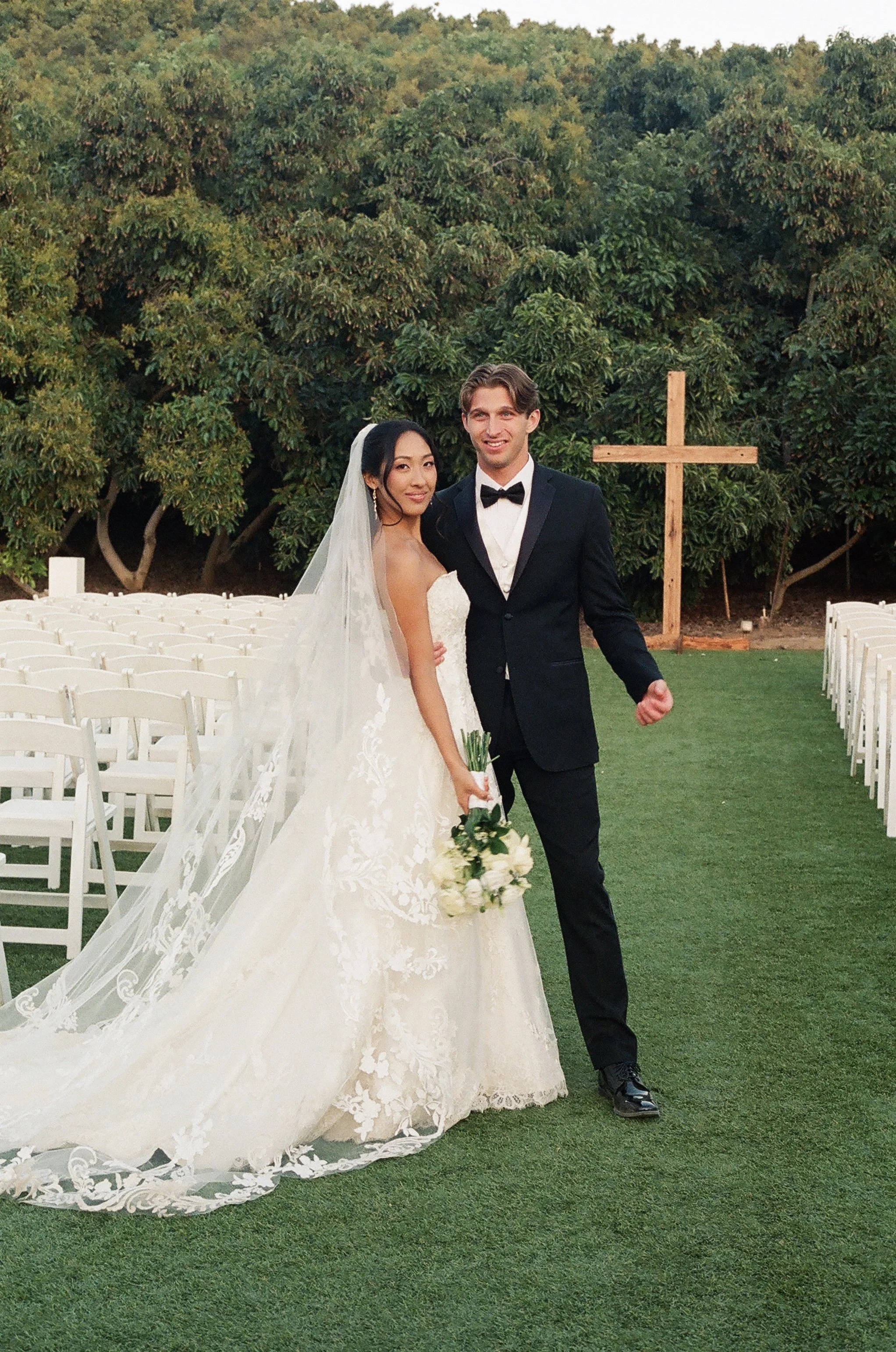 A bride and groom standing outdoors on a grassy area for a wedding ceremony, with a large wooden cross in the background, surrounded by trees.